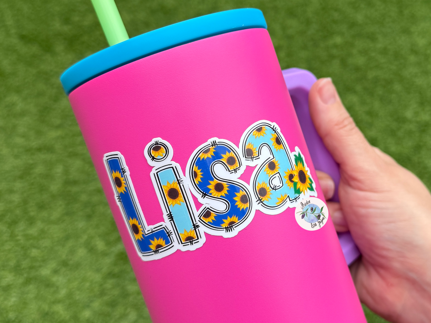 Pink tumbler with 'Lisa' and sunflower design sticker held by a hand on a grass background