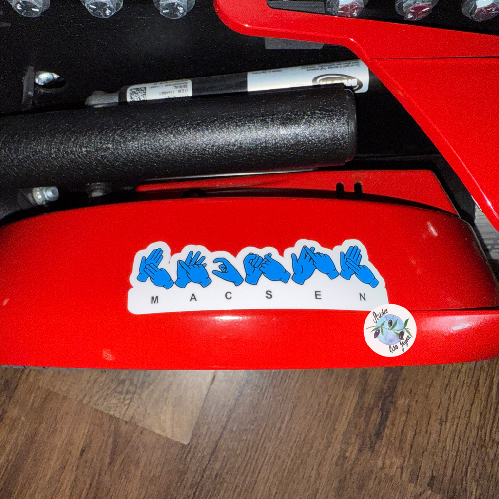 Close-up of a red electric wheelchair with a sticker featuring blue hand icons spelling Macsen in BSL (British Sign Language)