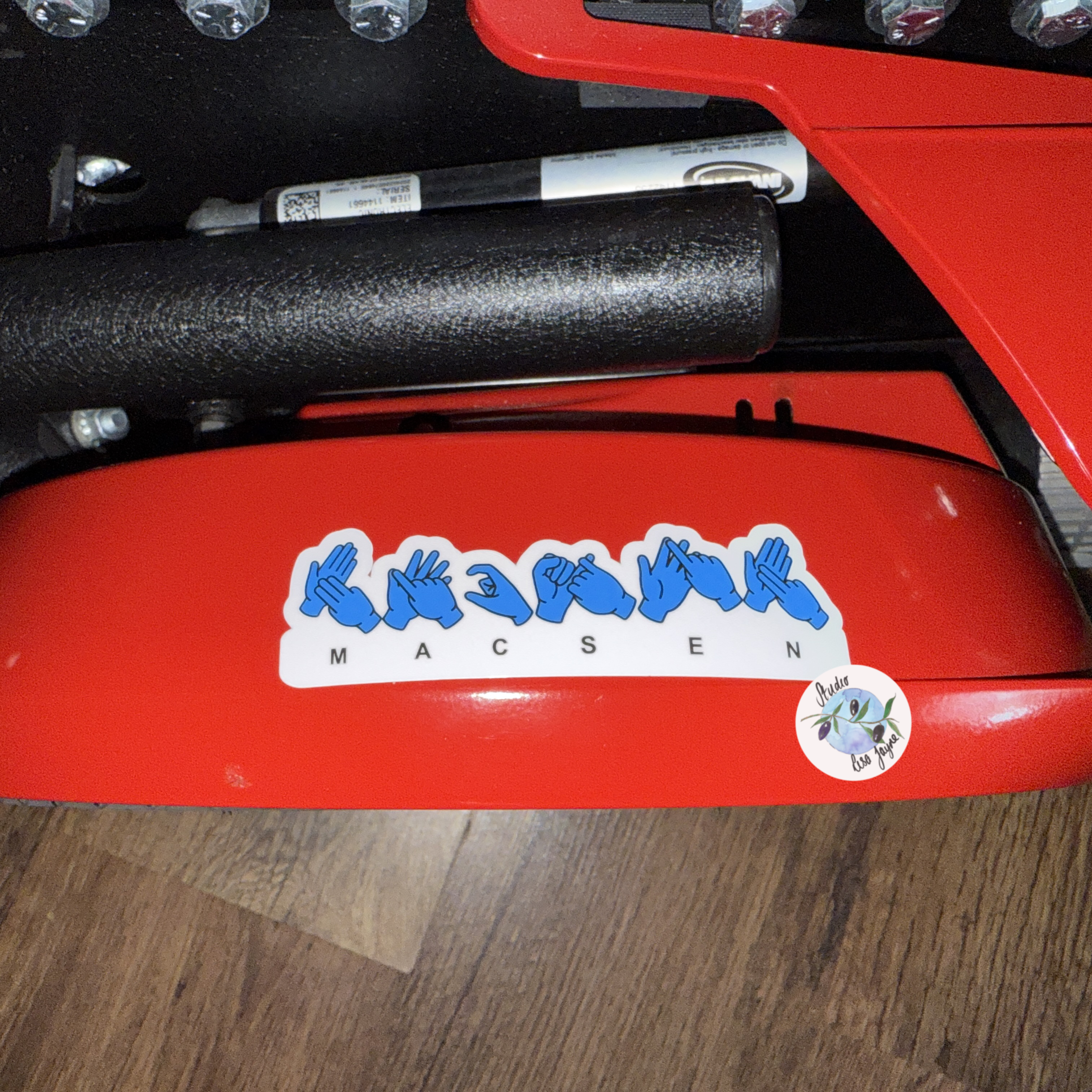 Close-up of a red electric wheelchair with a sticker featuring blue hand icons spelling Macsen in BSL (British Sign Language)