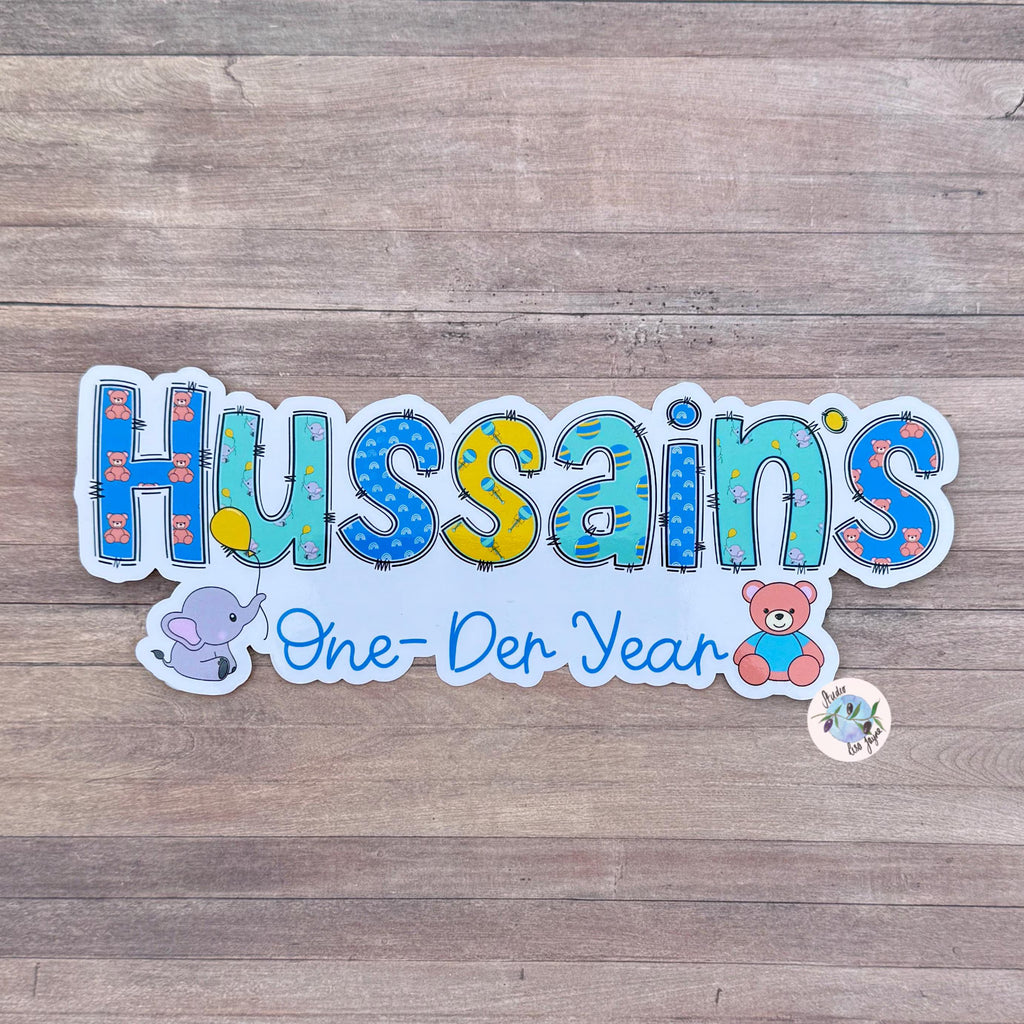 Colorful new baby themed sticker with 'Hussains' One Der Year' text on a wooden surface.  Brights colour style