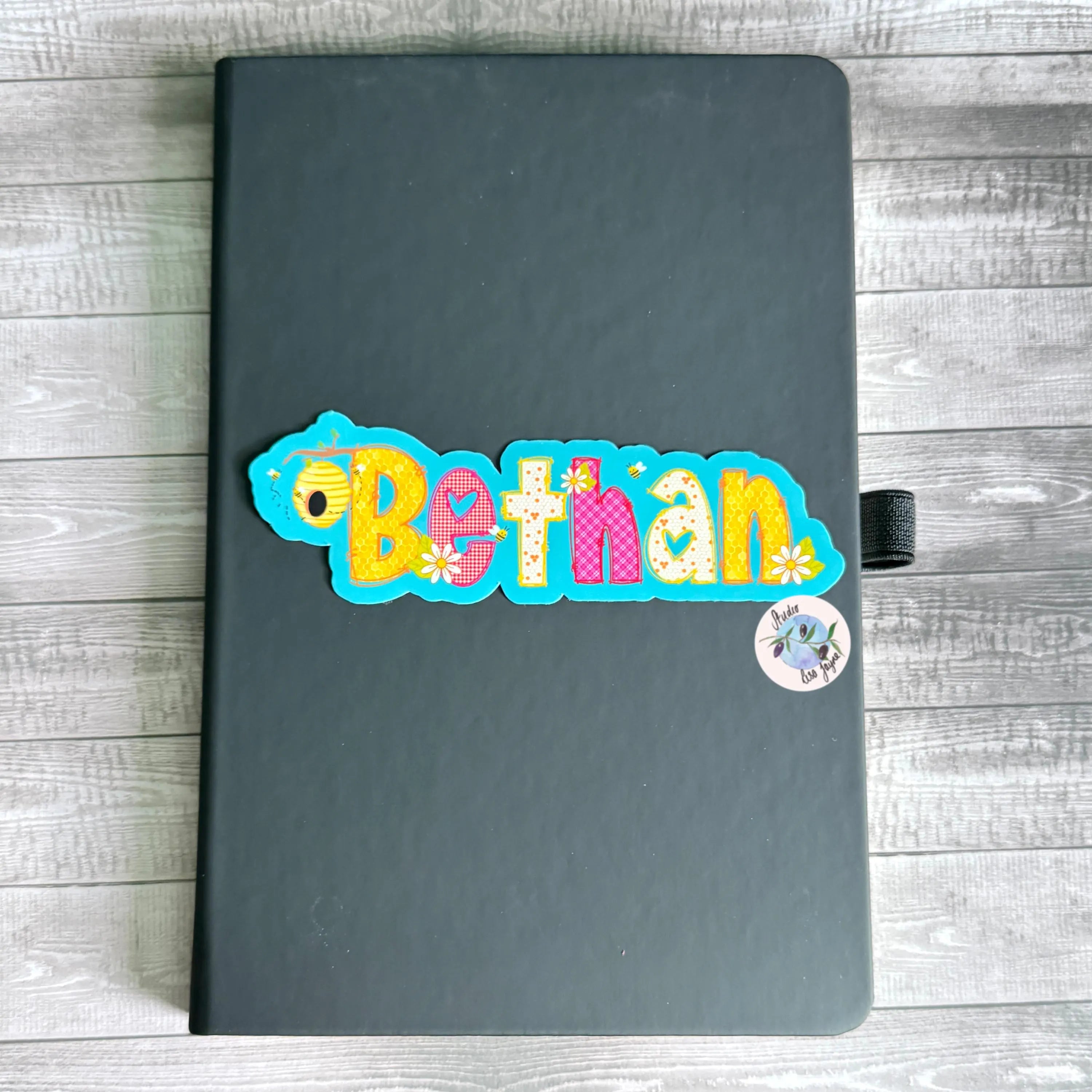 Black notebook diary journal with a colorful bee themed 'Bethan' sticker on a wooden surface.  BLue Pink Yellow