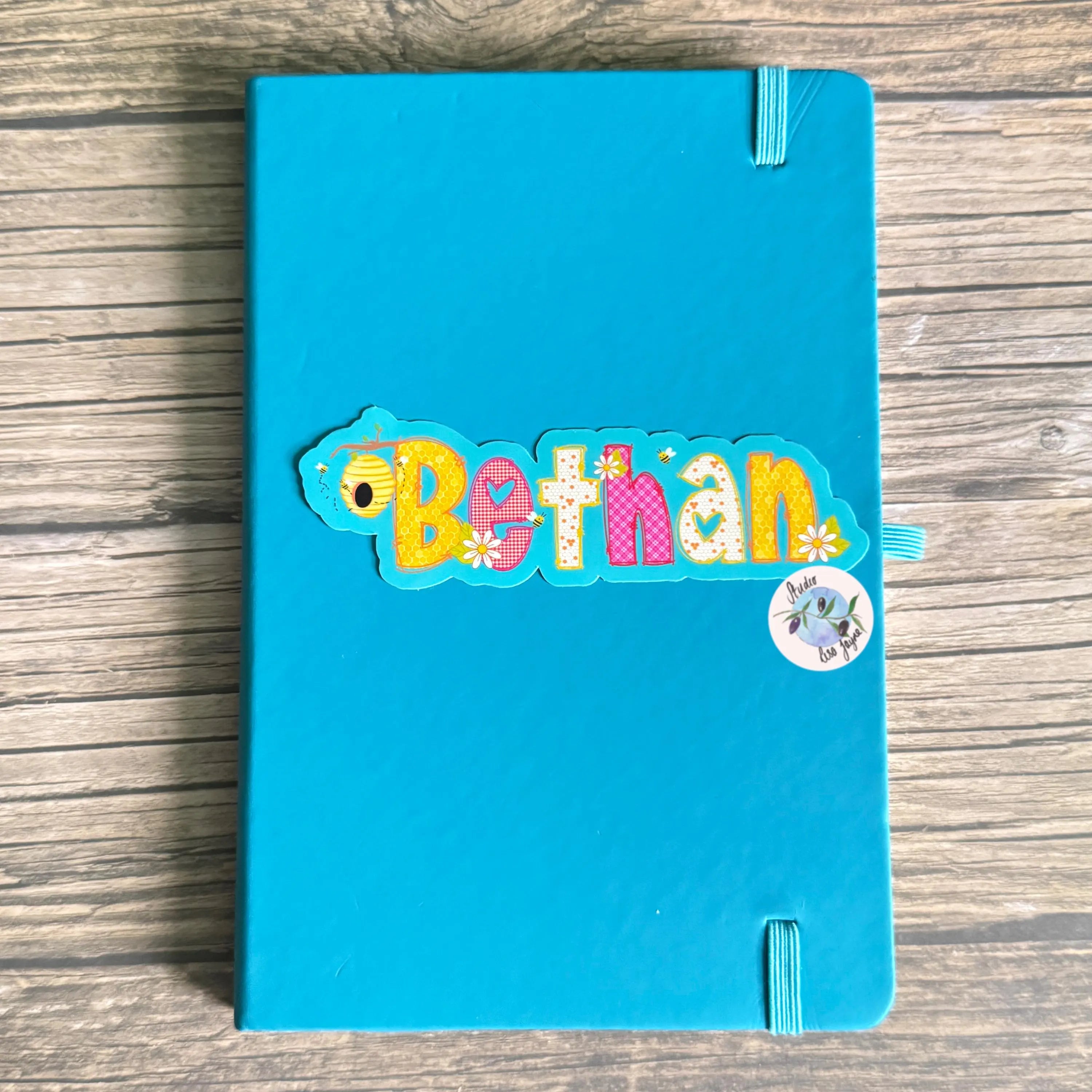 Blue notebook with colorful summer name 'Bethan' sticker on a wooden surface