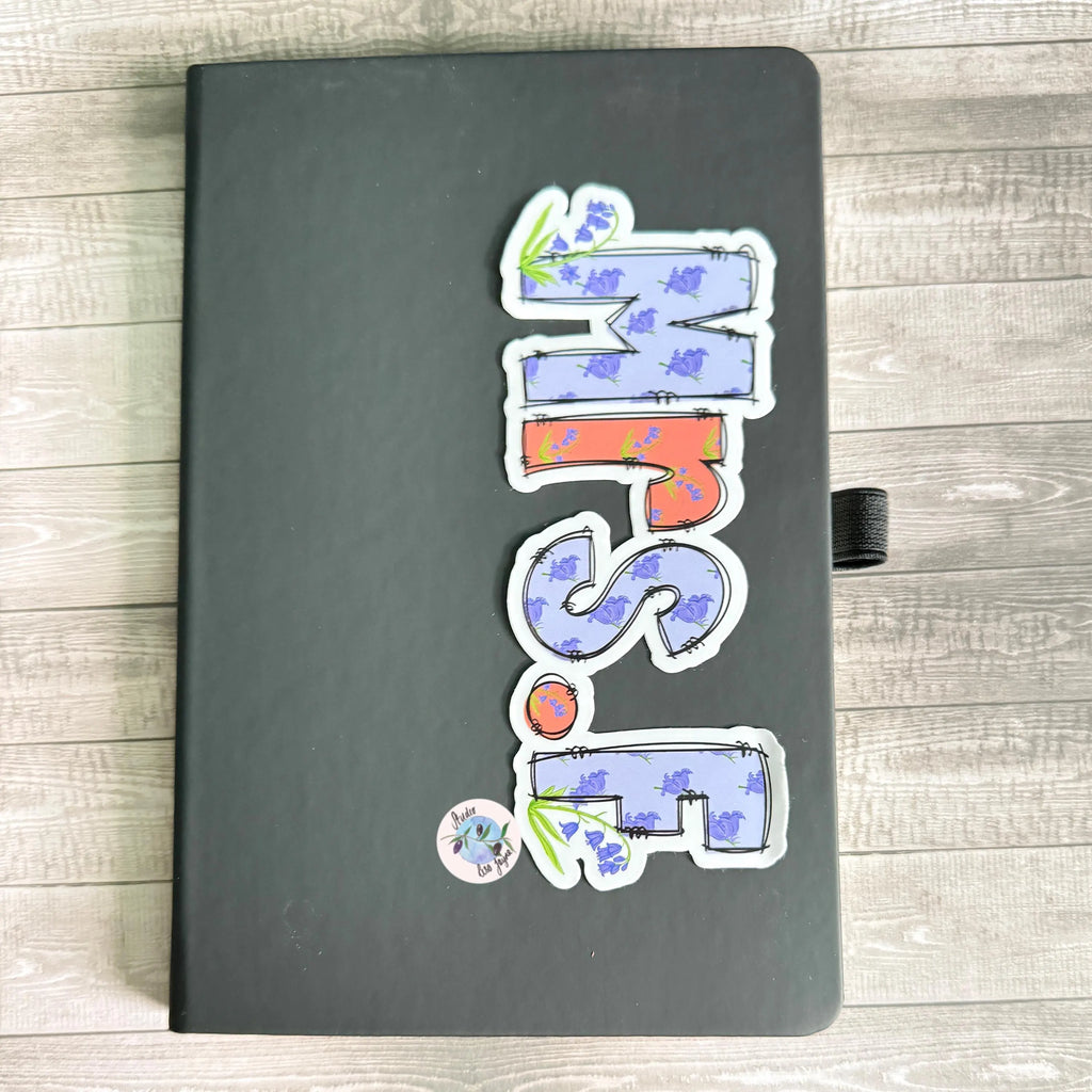 Black notebook with colorful stickers spelling 'Mrs F' on a wooden surface