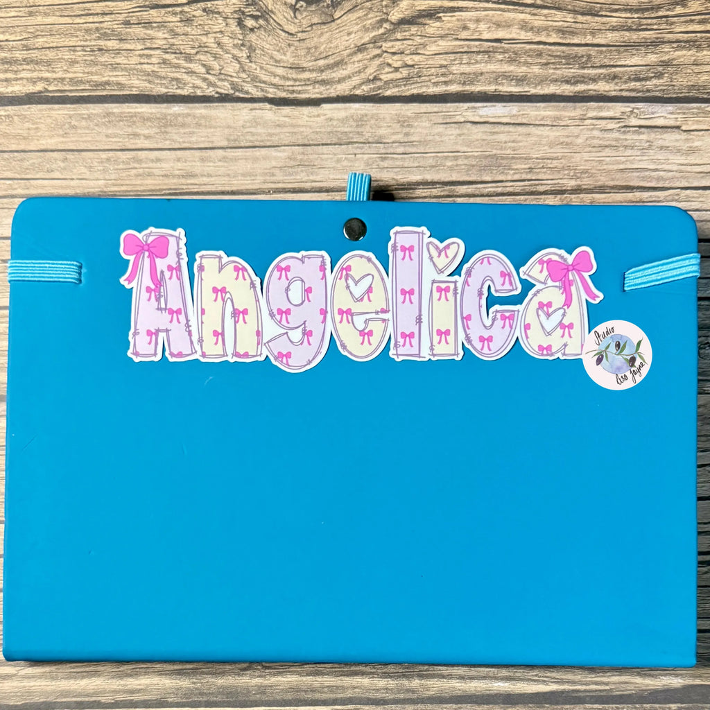 Blue journal notebook diary with 'Angelica' written on it, featuring decorative pink bows, on a wooden surface.