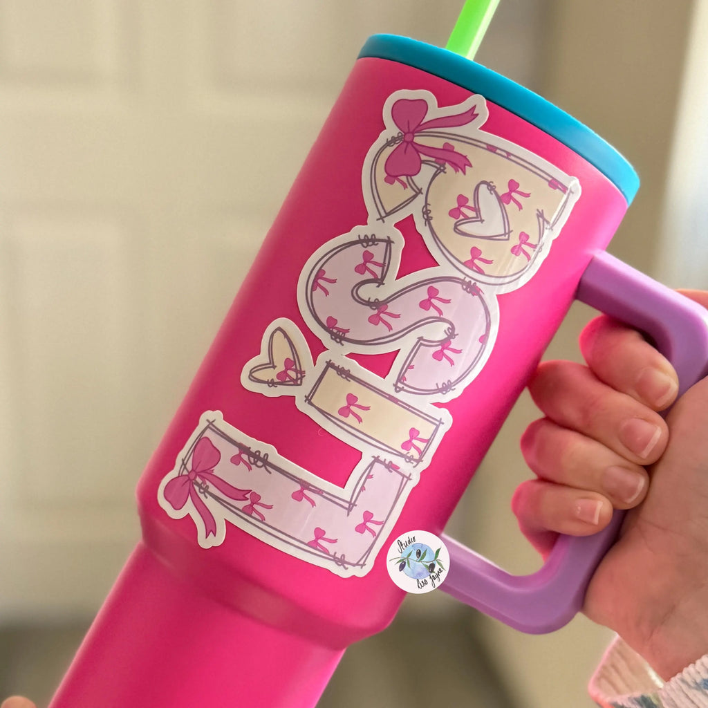 Pink sippy cup with decorative pink bow name sticker and a handle, held by a hand against a neutral background.