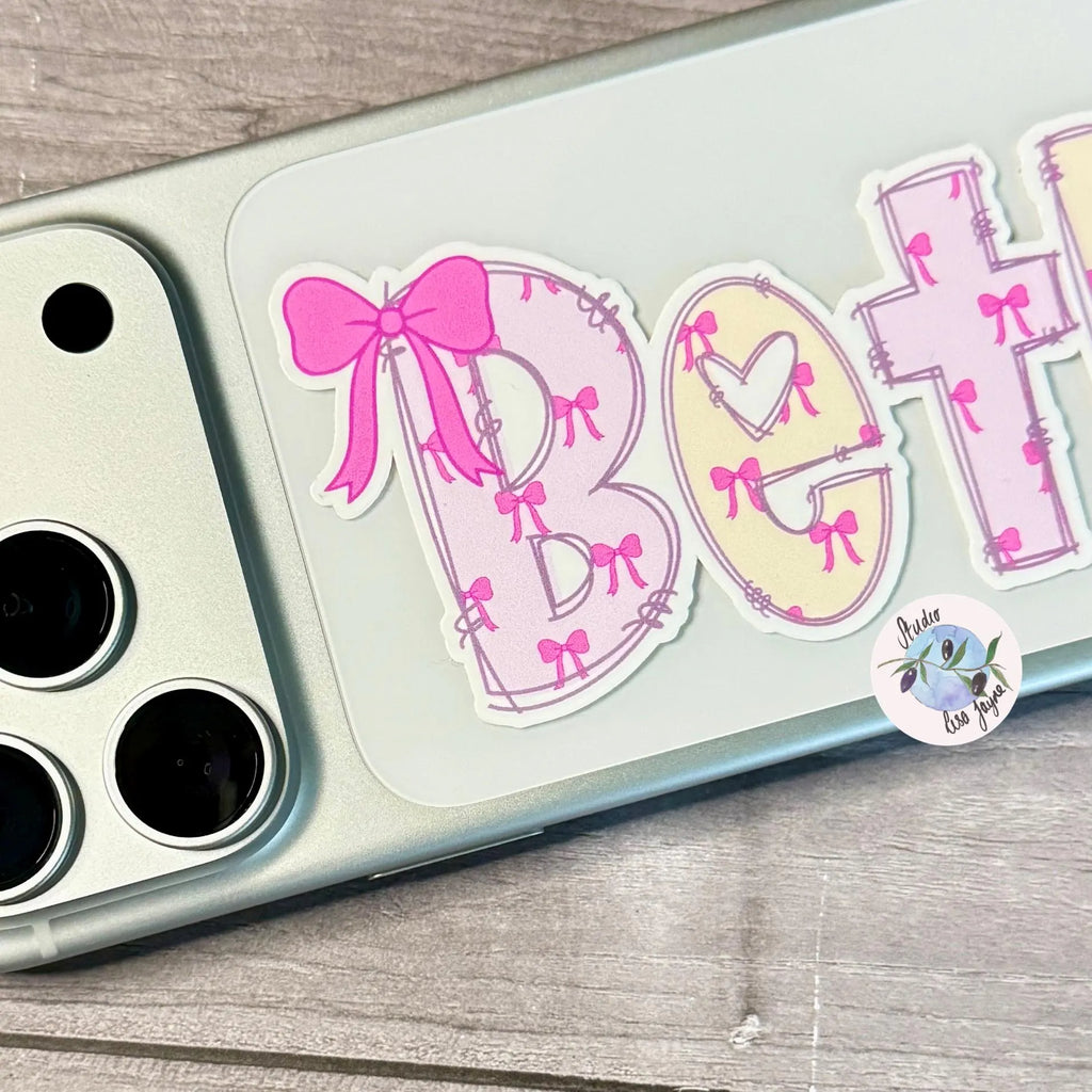 Phone case with decorative stickers featuring letters and a pink bow on a wooden surface