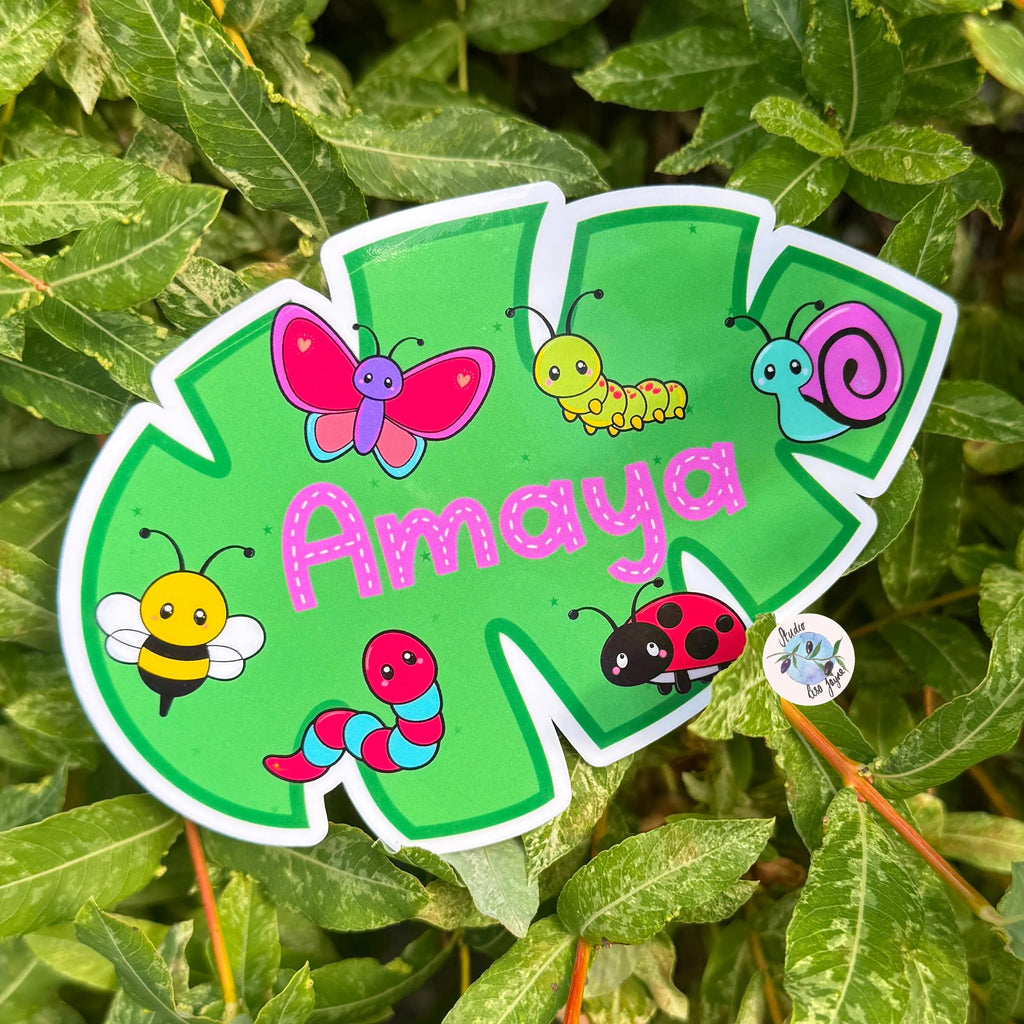 Green leaf shaped name sticker with cartoon insects and pink name 'Amaya' on a leafy background