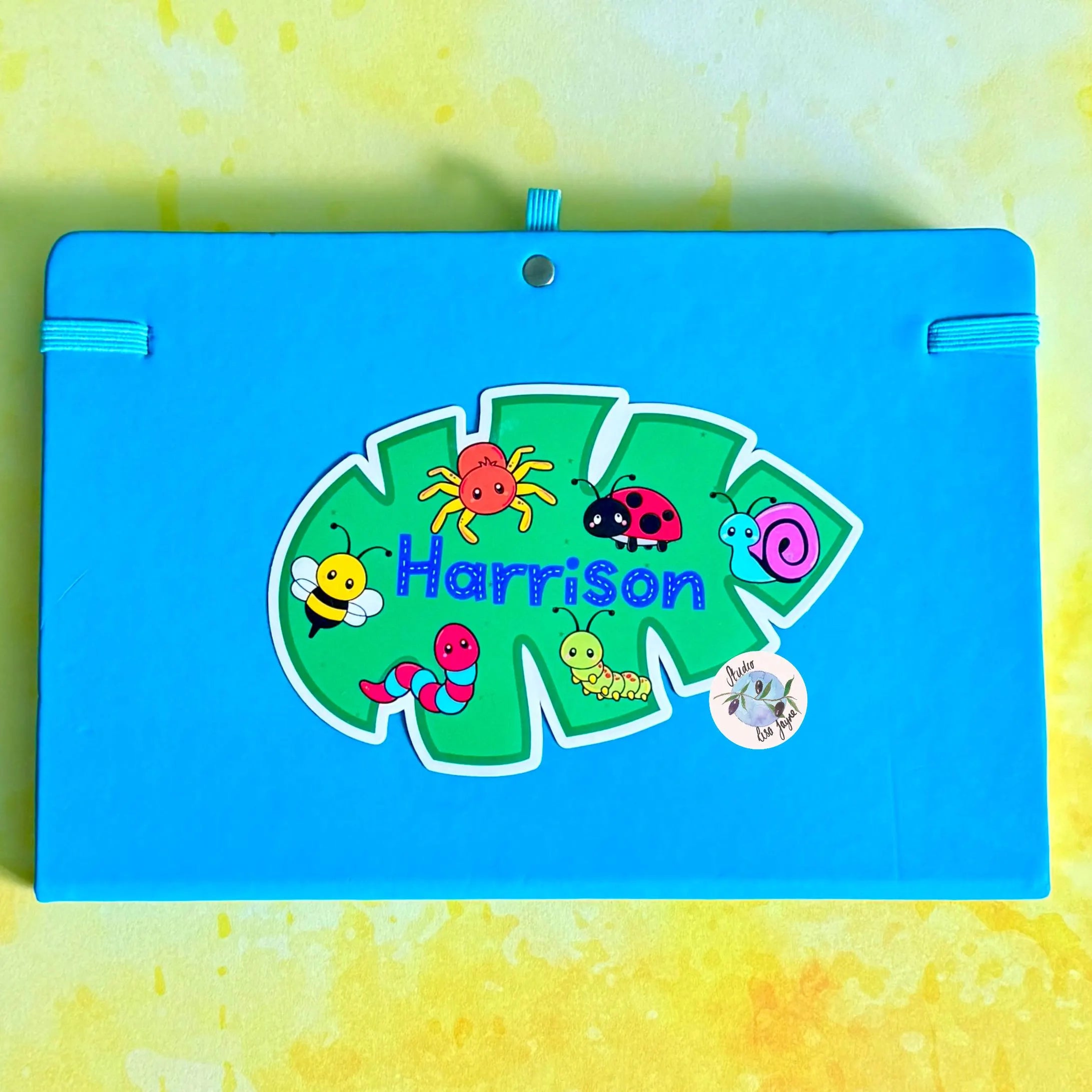 Blue notebook diary with colorful insect illustrations and the name 'Harrison' on a yellow and green textured background.