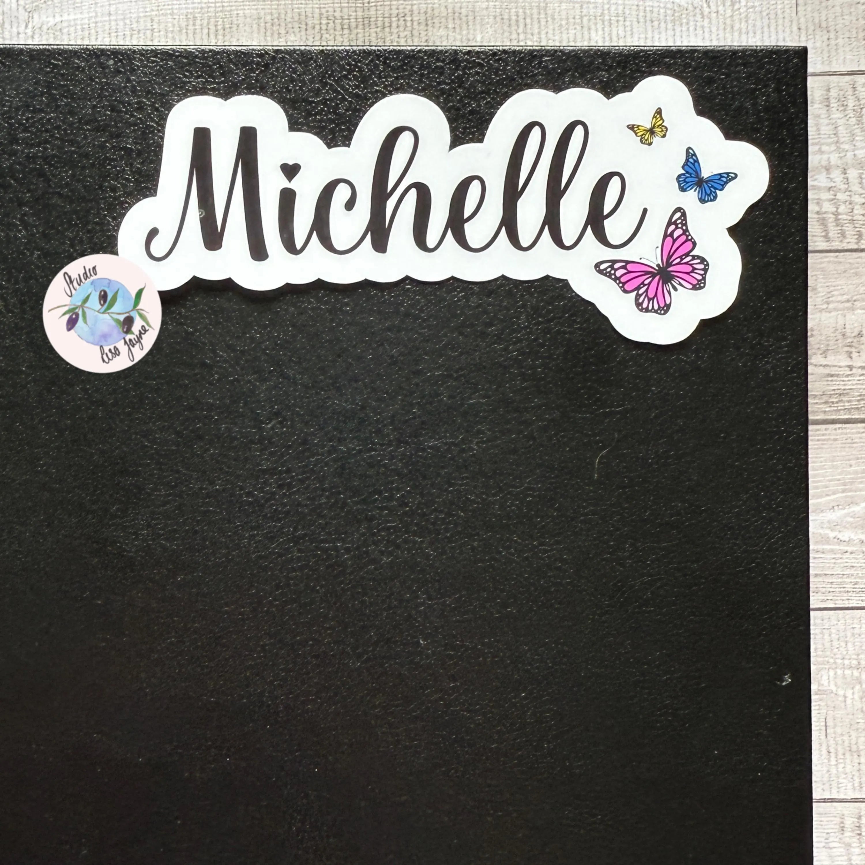 Sticker with 'Michelle' and pink, yellow and blue butterflies on a black diary notebook journal
