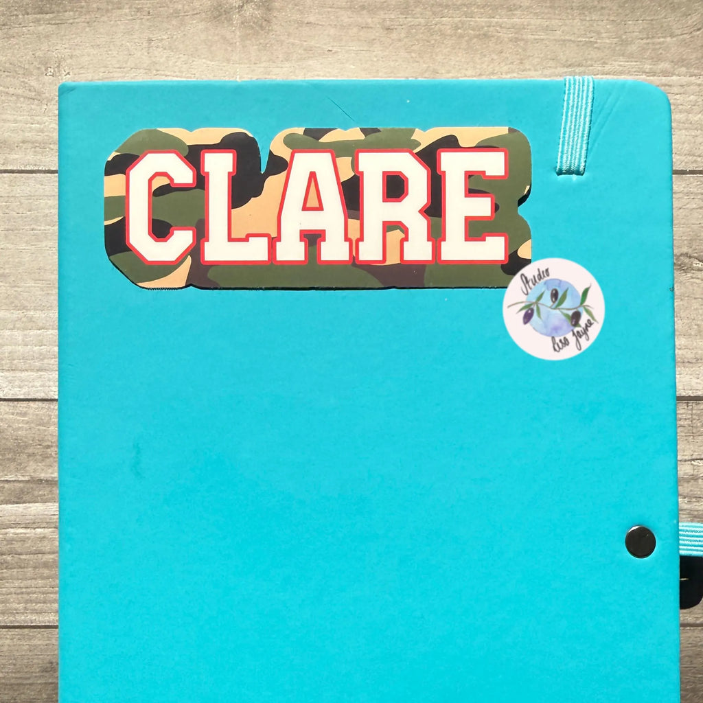 Turquoise notebook with 'CLARE' camouflage sticker on a wooden surface