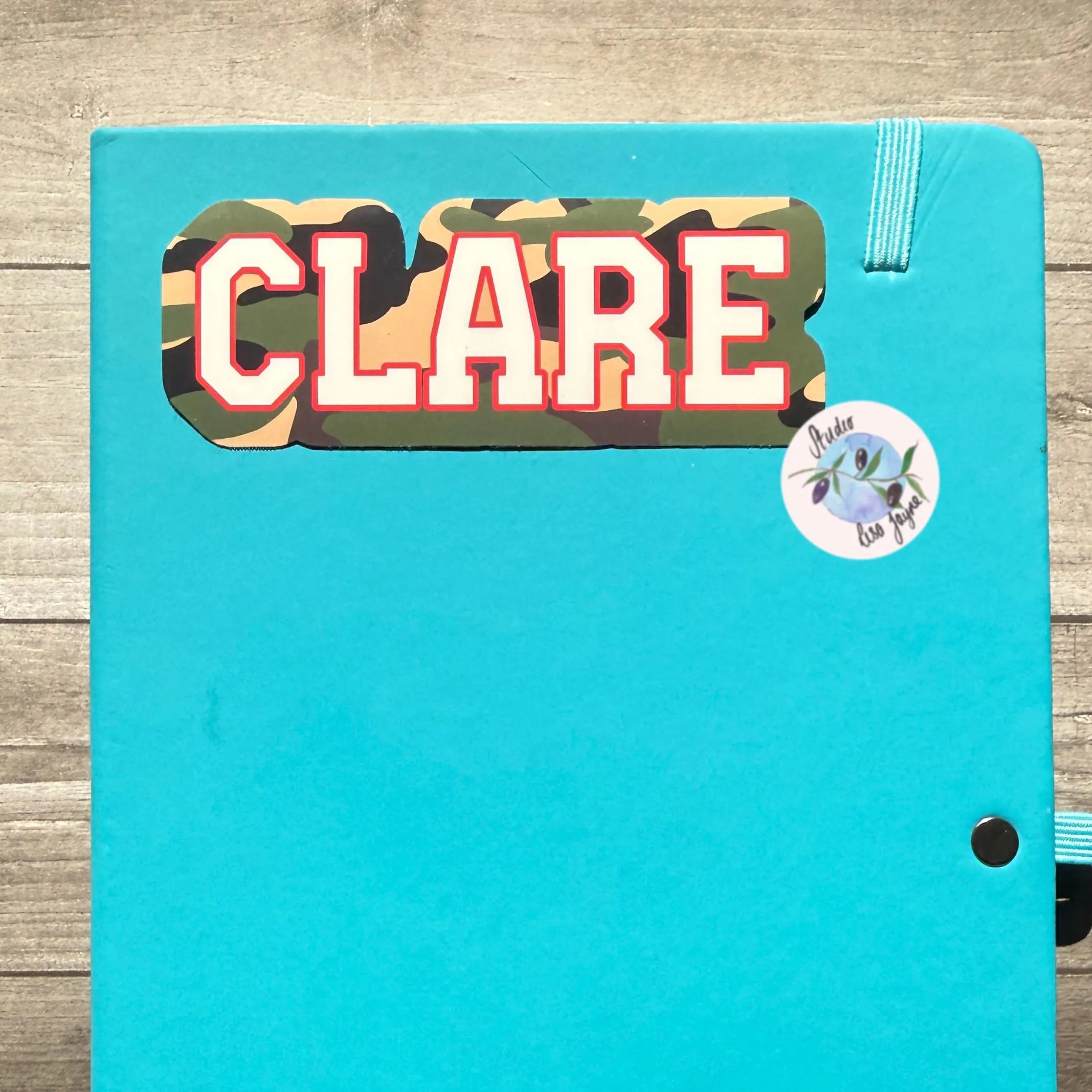 Turquoise notebook with 'CLARE' camouflage sticker on a wooden surface