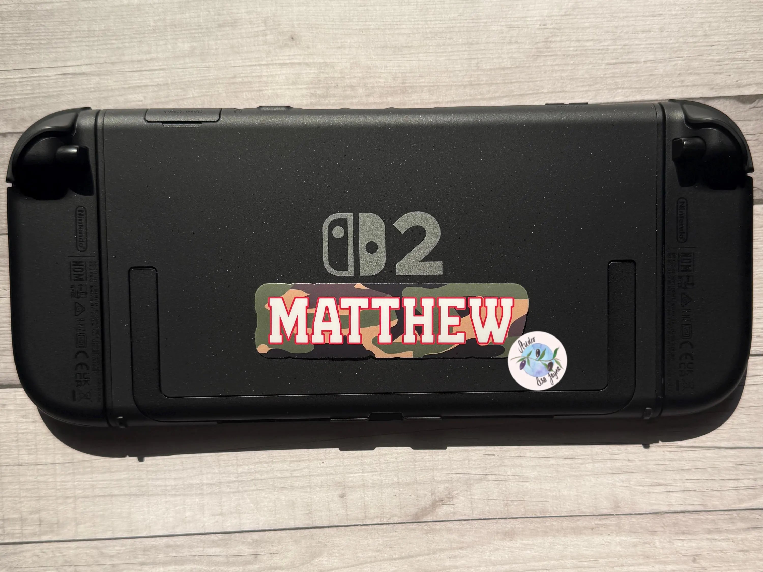 Black gaming console with 'Matthew' camouflage sticker on a wooden surface.  Nintendo Switch 2