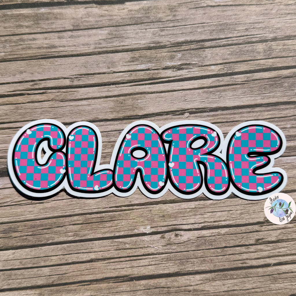 Checkered name sticker 'CLARE' on a wooden surface