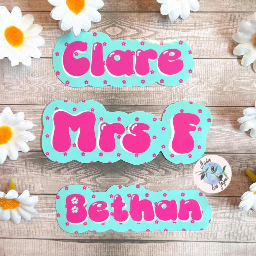 Colorful name labels with 'Clare', 'Mrs F', and 'Bethan' in a cherry blossom design  on a wooden surface with daisies.