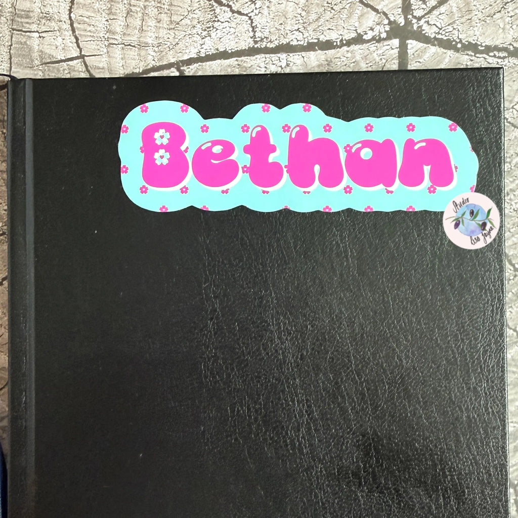 Black book with a decorative sticker spelling 'Bethan' on a textured surface