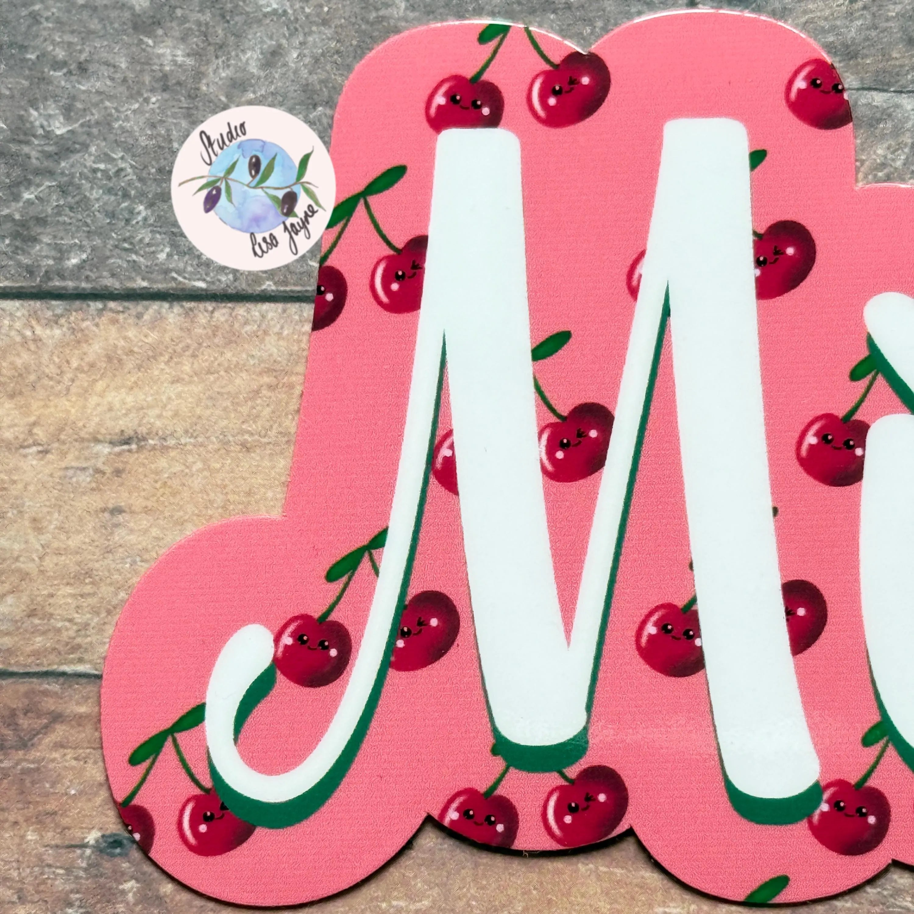 Pink letter 'M' with cherry design on a wooden surface
