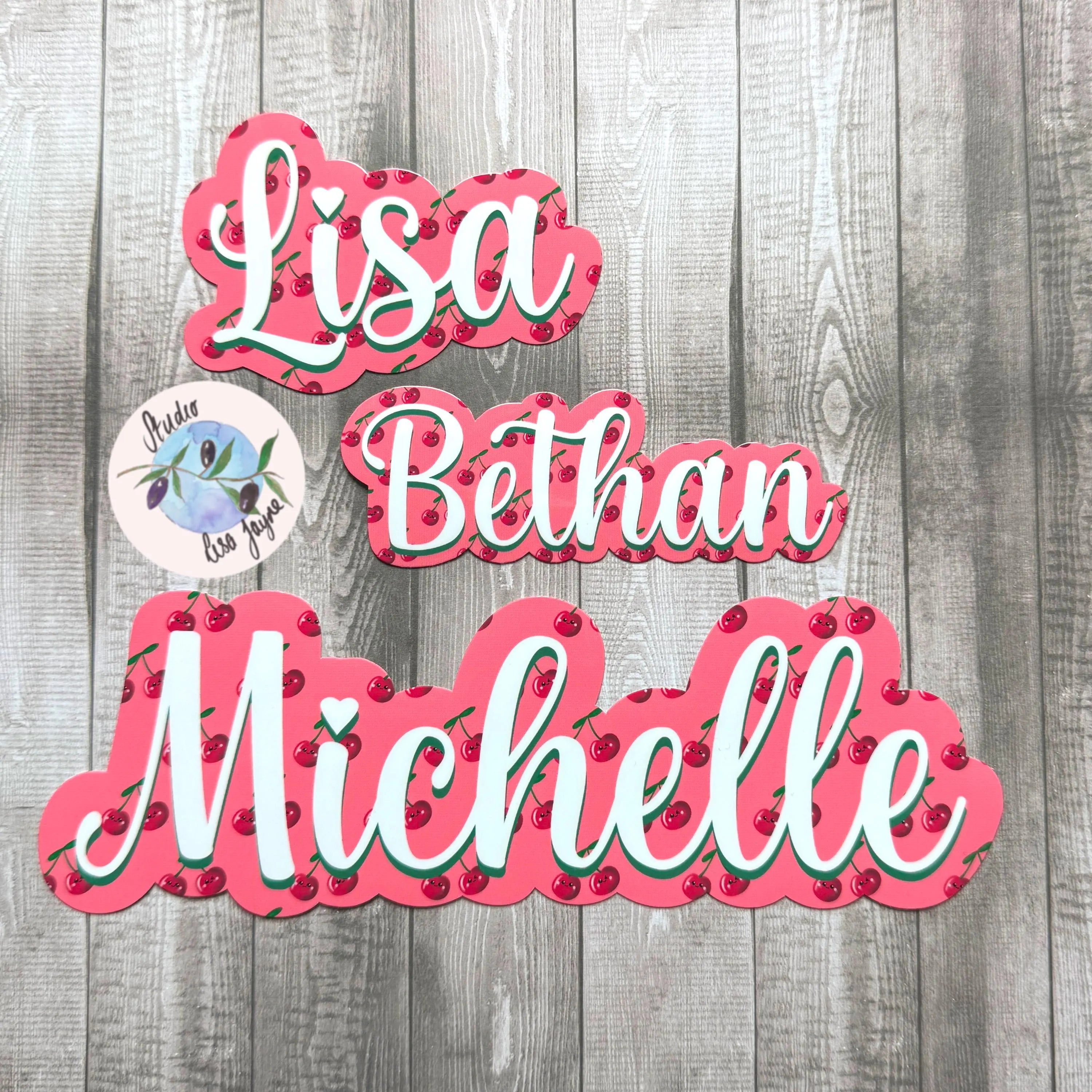 Three decorative cherry name stickers with 'Lisa', 'Bethan', and 'Michelle' on a wooden background.