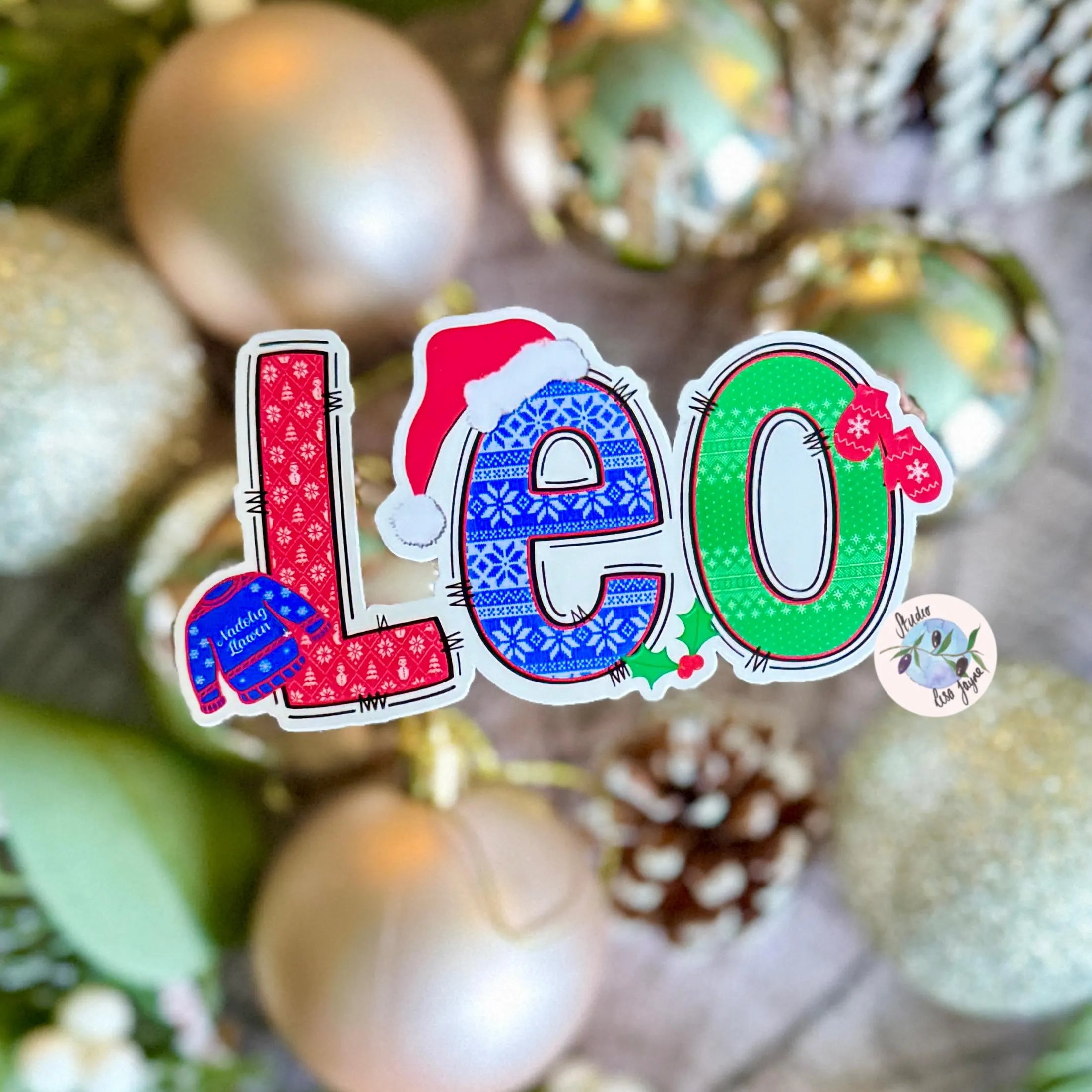 Colorful 'Leo' Christmas Jumper name sticker on a blurred background of ornaments.
