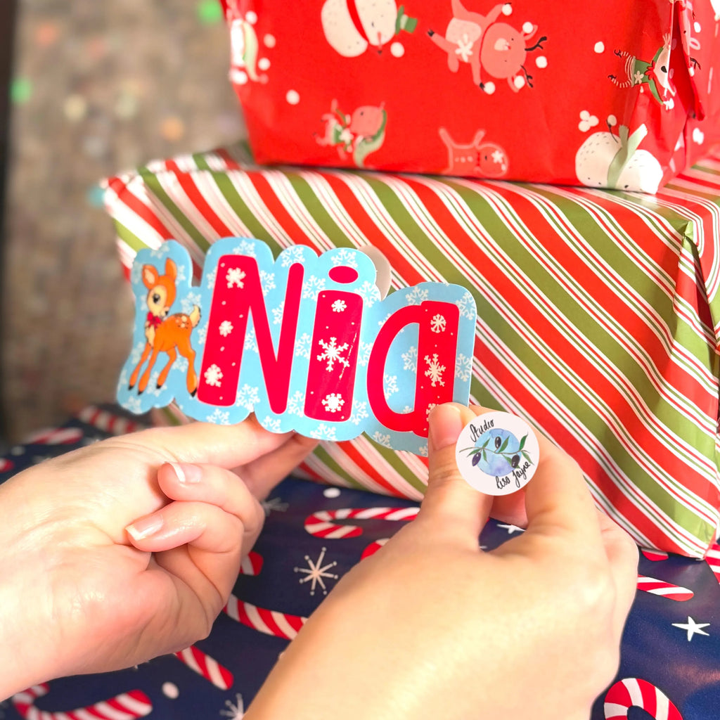 Person holding a decorative name tag with 'Nia' and a deer, with Christmas-themed wrapped gifts in the background.