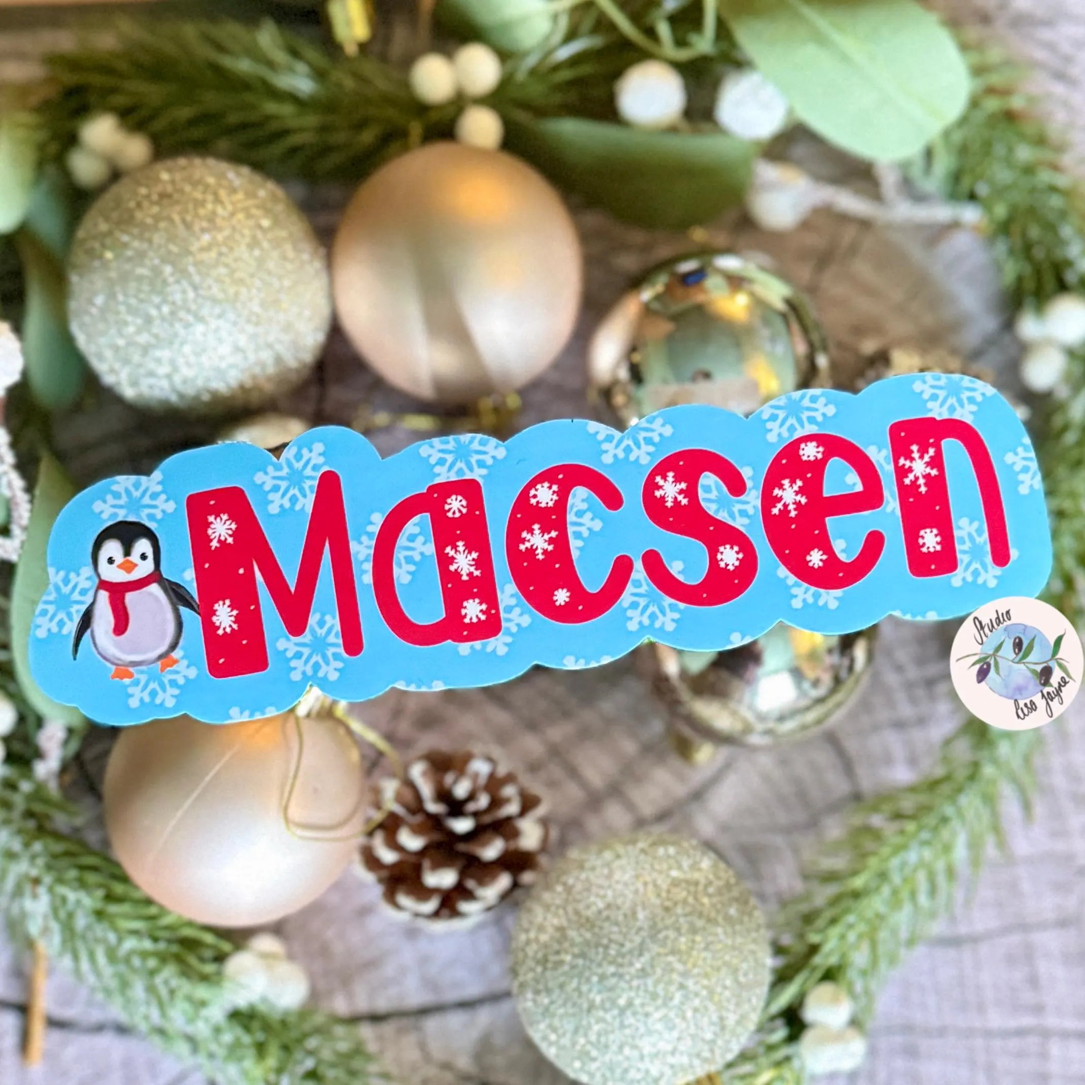 Decorative personalised name sticker with 'Macsen' and a penguin surrounded by Christmas ornaments and greenery