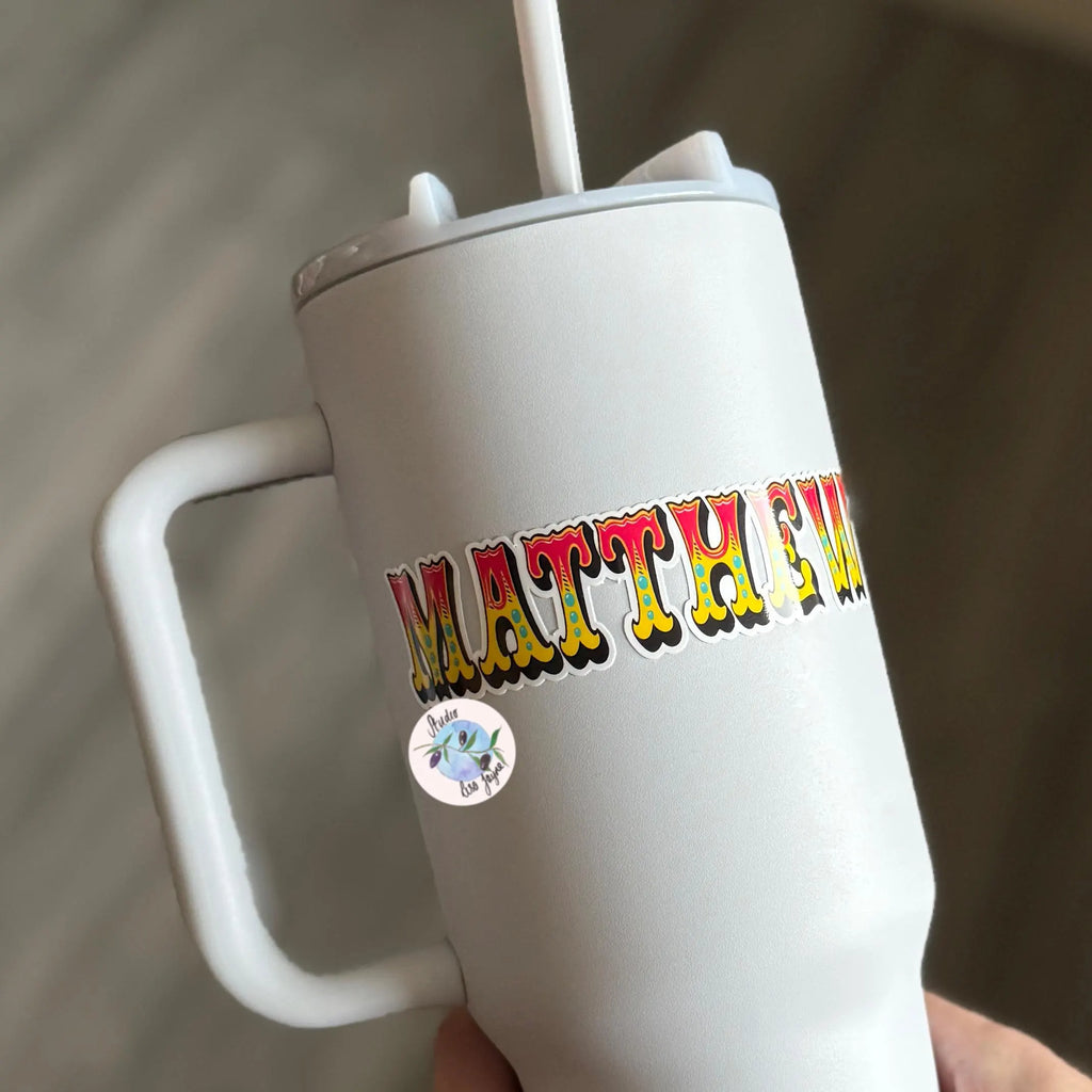 White tumbler with colorful circus text 'MATTHEW' vinyl decal held by a hand against a neutral background