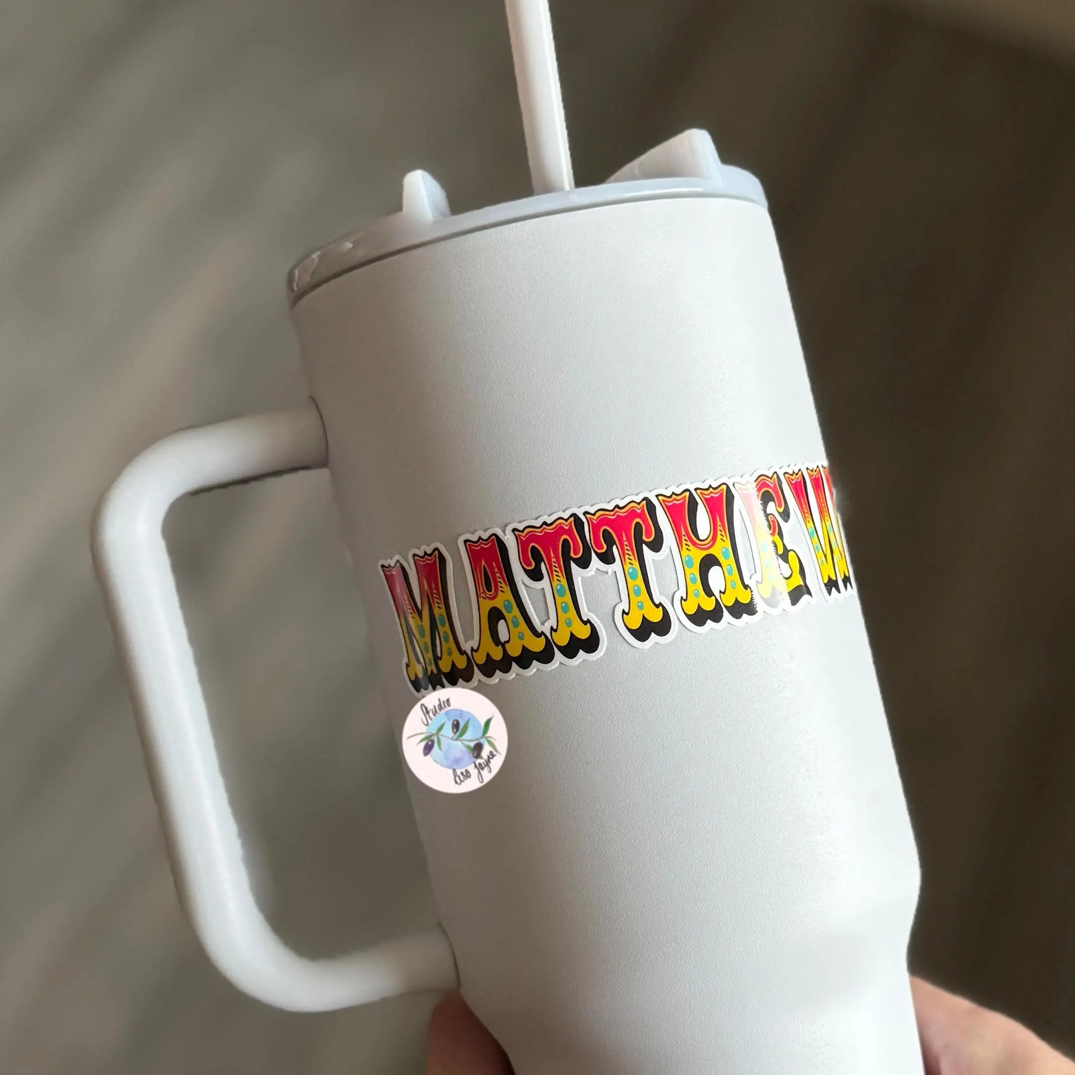 White tumbler with colorful circus text 'MATTHEW' vinyl decal held by a hand against a neutral background