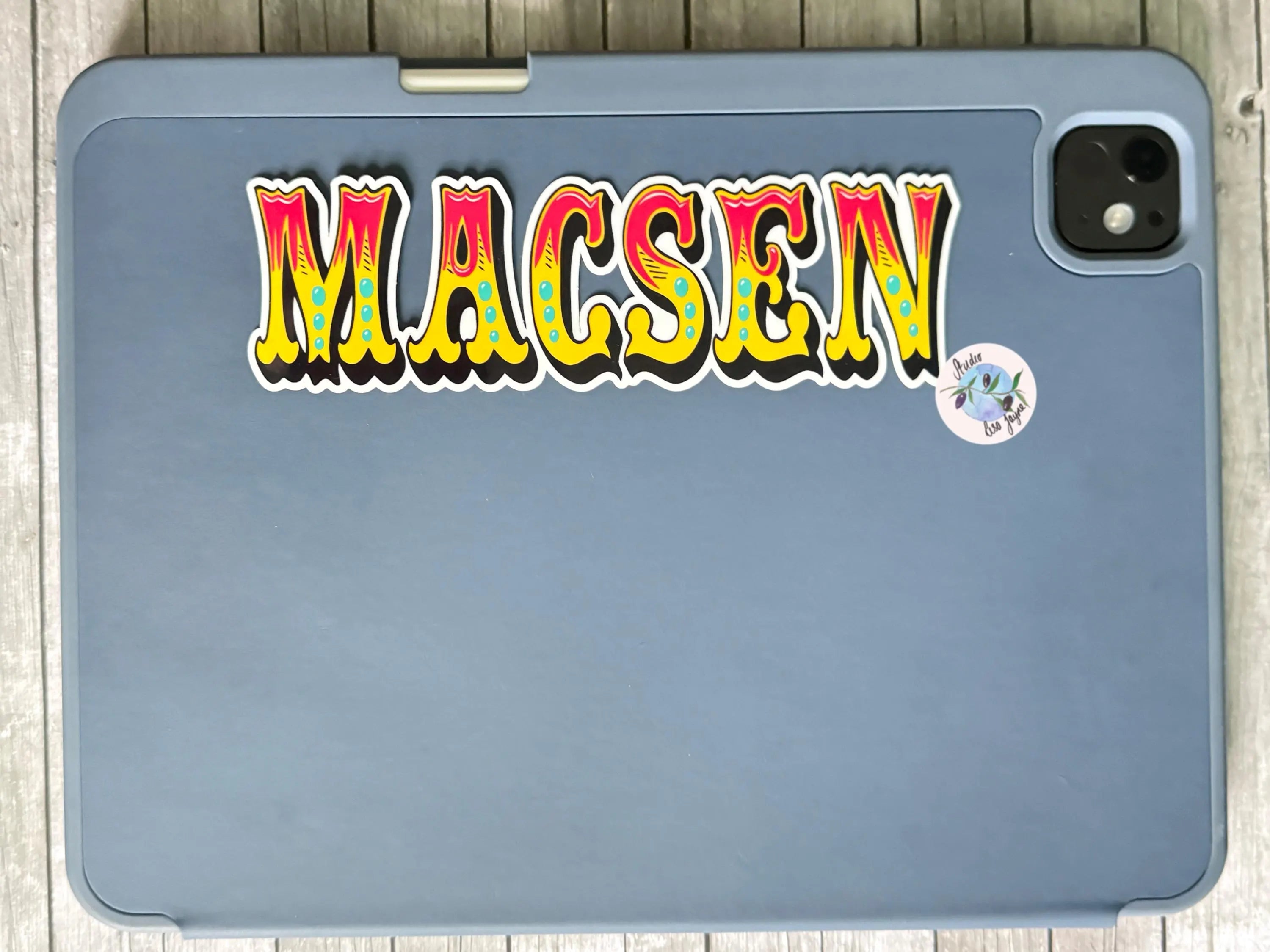 Blue tablet case with colorful 'MACSEN' old style font sticker on a wooden surface