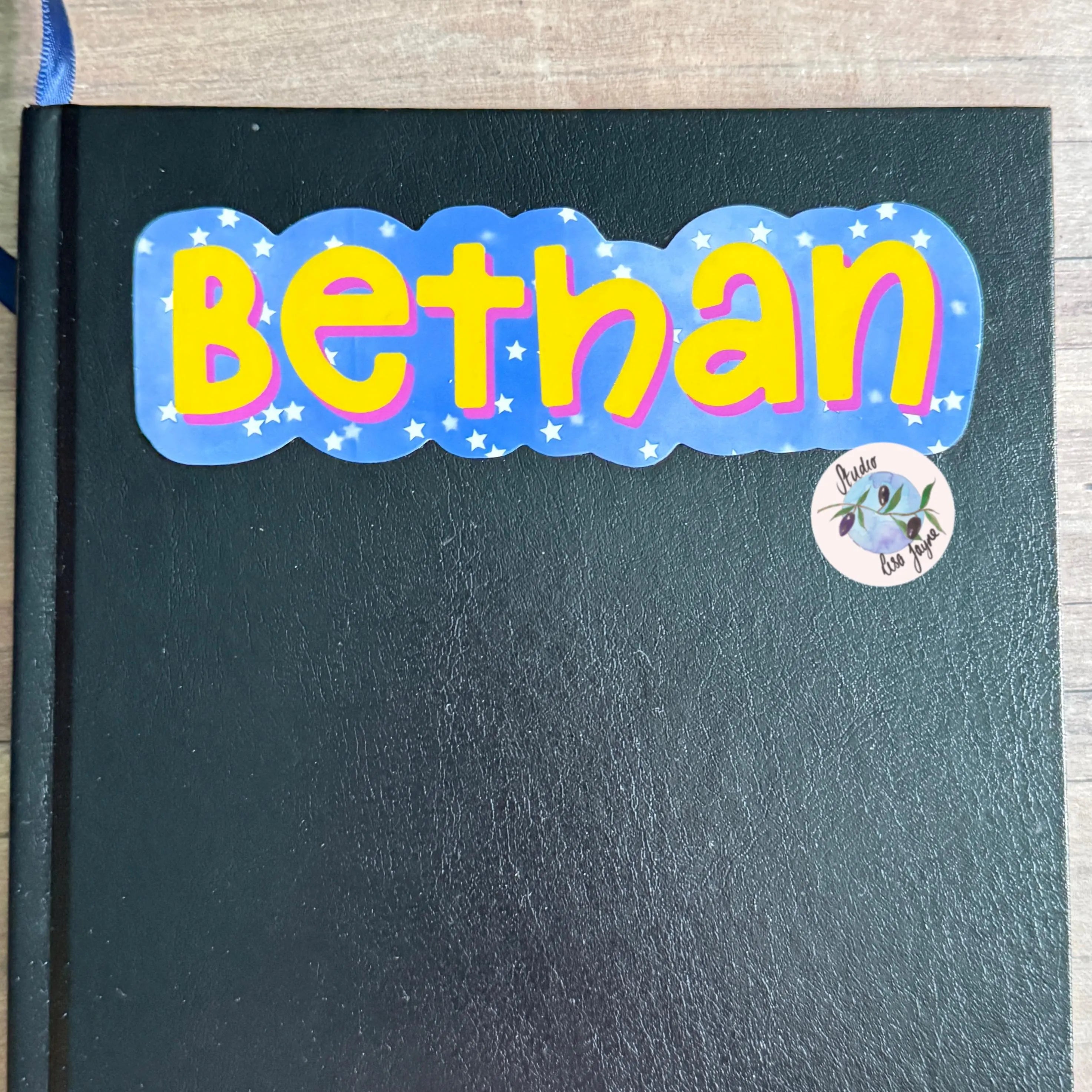 Black notebook with a colorful star themed cute name sticker reading 'Bethan'.  Colours are blue, purple, pink and yellow