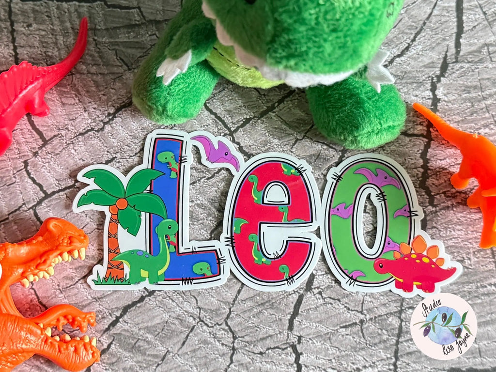 Sticker with colorful letters spelling 'LEO' surrounded by toy dinosaurs on a textured surface