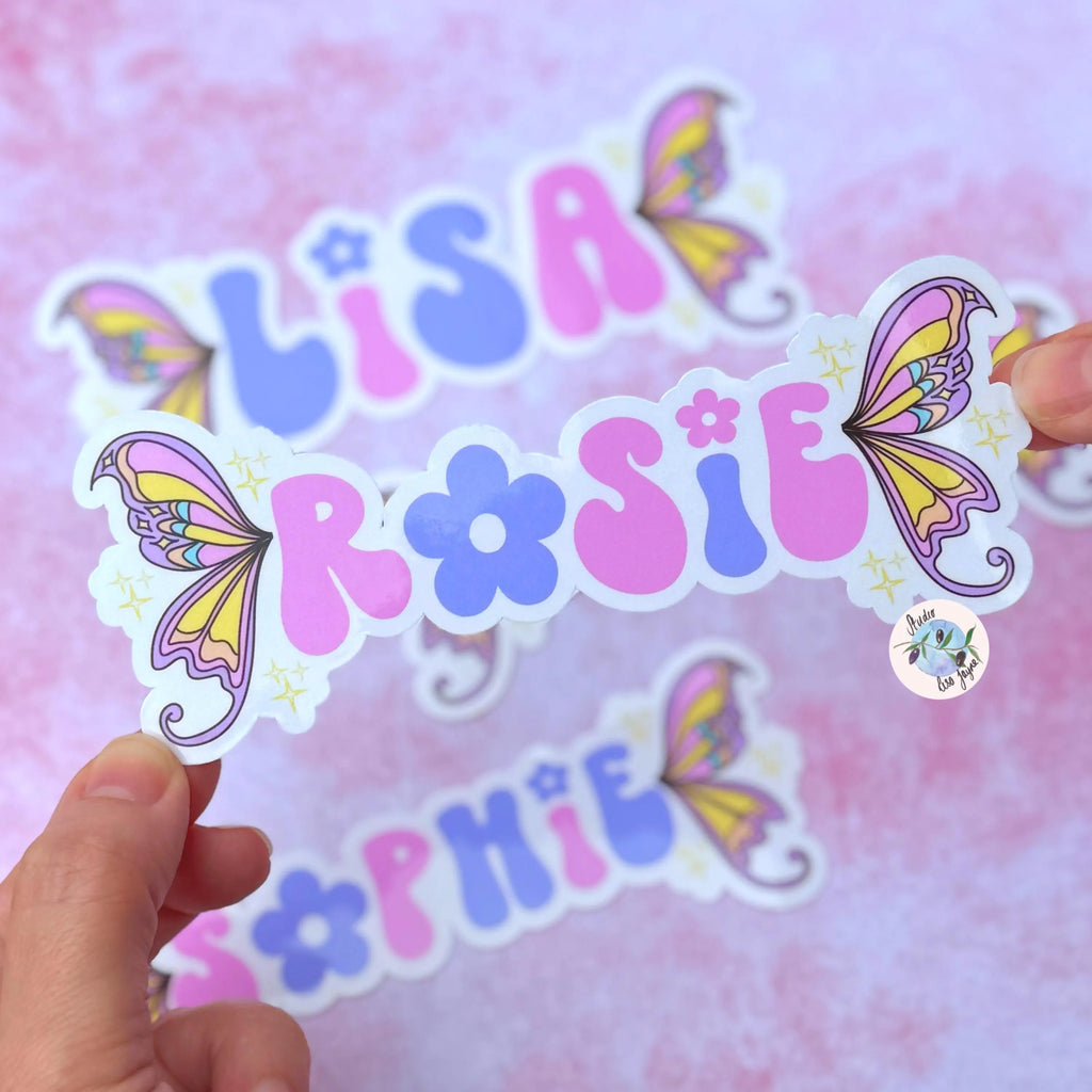 Colorful name stickers with fairy wing design held against a pink and purple background