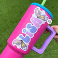 Pink tumbler with 'Lisa' and fairy wing designs on a green background
