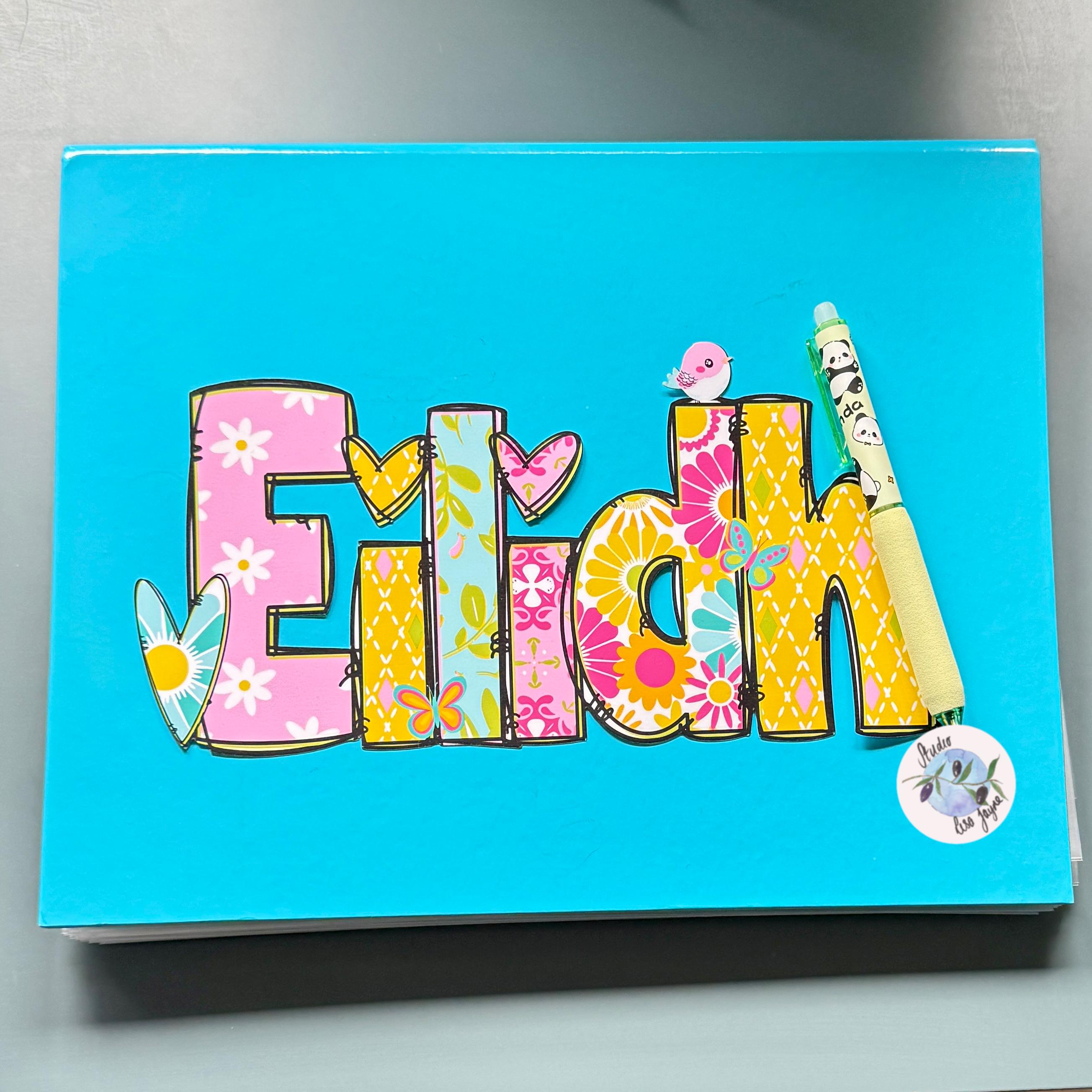 A bright turquoise notebook or journal cover featuring the name 'Eilidh' in large decorative letters with floral and patterned fills, plus small bird and butterfly illustrations, photographed on a neutral gray surface.