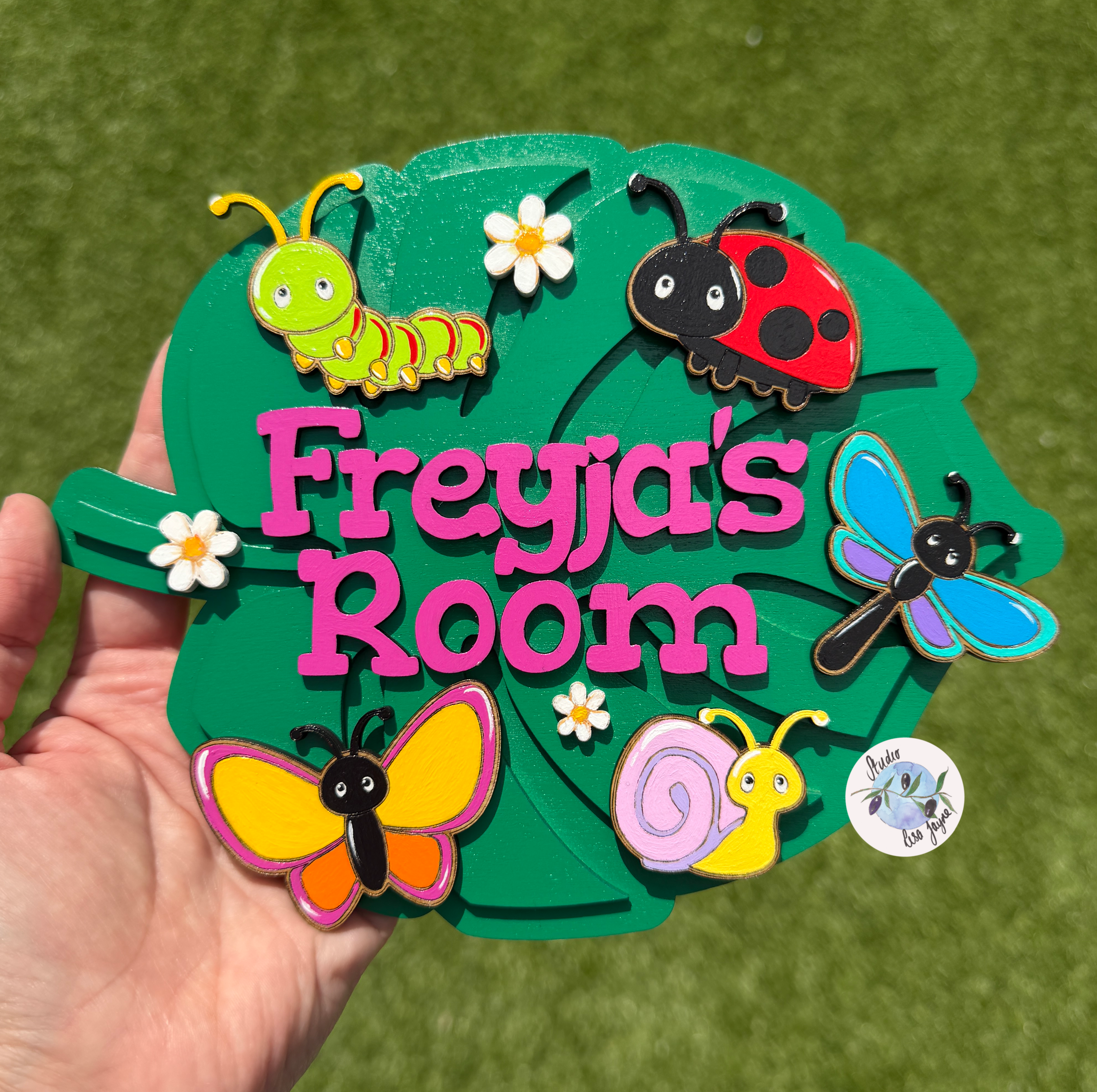 A hand holds a green leaf-shaped wooden sign that reads 'Freyja's Room' in pink letters, decorated with colorful nature-themed stickers including a ladybug, butterfly, snail, caterpillar, dragonfly, and small flowers. The background is green grass.