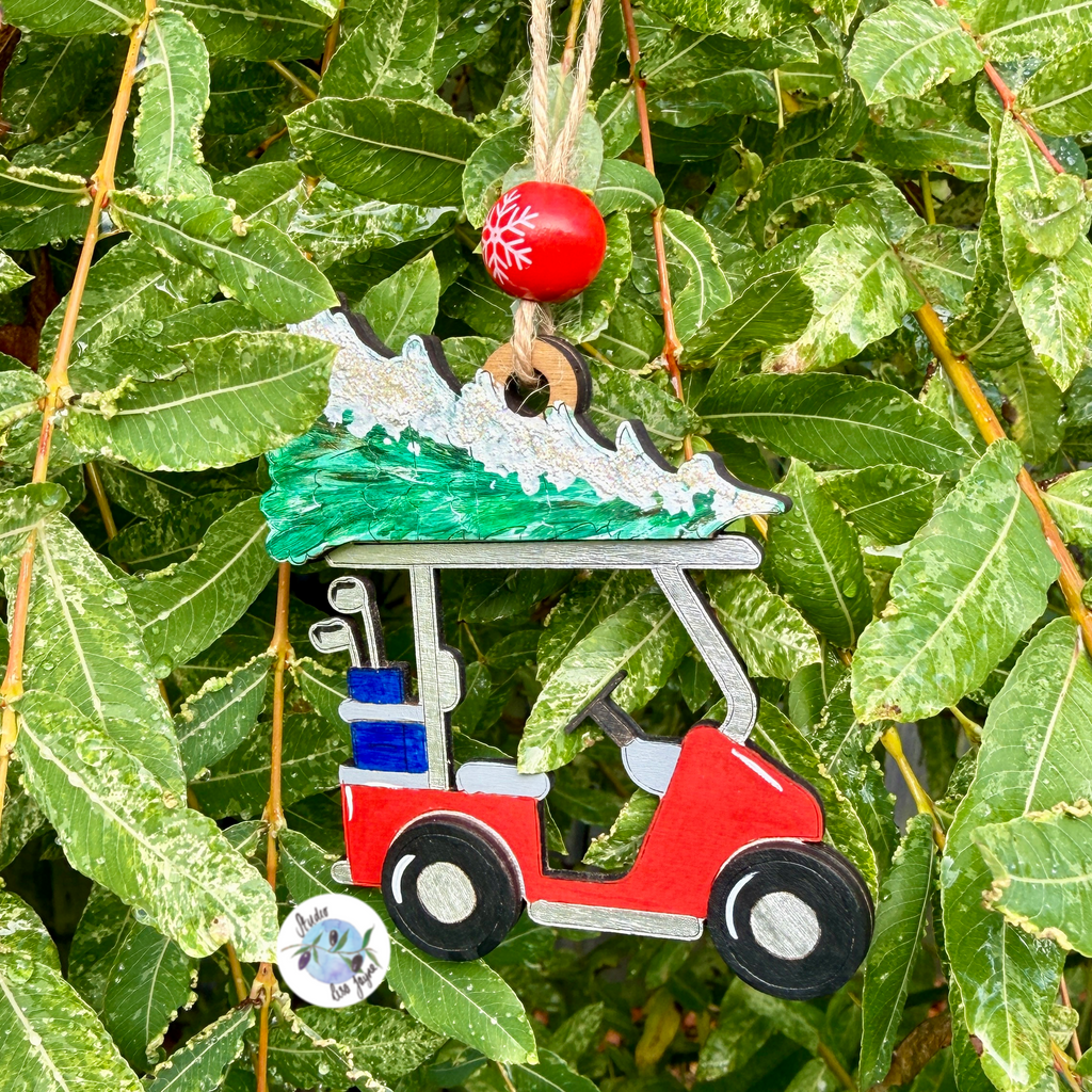 Handpainted decorative ornament of a golf cart with Christmas tree and clubs, hanging from green leaves.