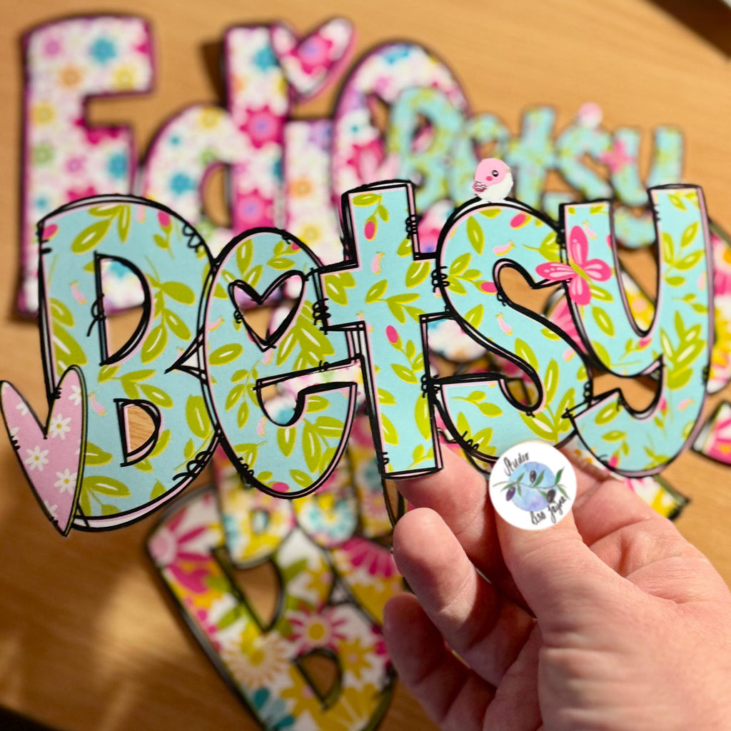 A hand holds a colourful floral name sticker with the name Betsy in a green floral style with a pink heart and pink butterfly
