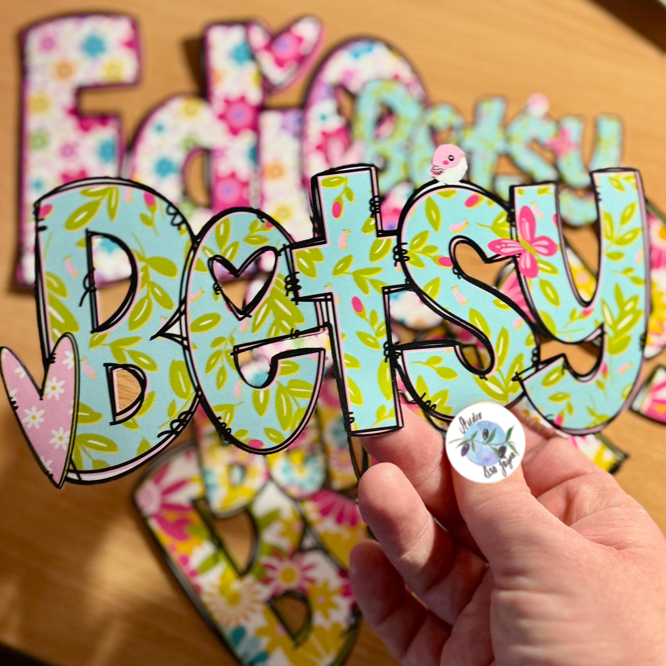 A hand holds a colourful floral name sticker with the name Betsy in a green floral style with a pink heart and pink butterfly