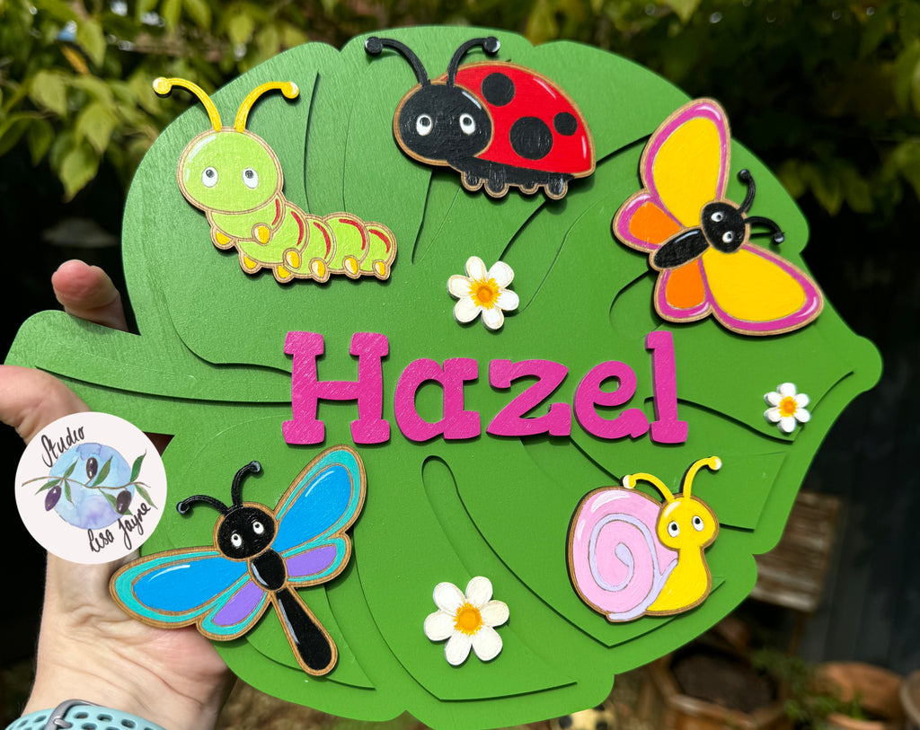 Decorative leaf-shaped sign with cartoon insects and flowers, featuring the name 'Hazel'.