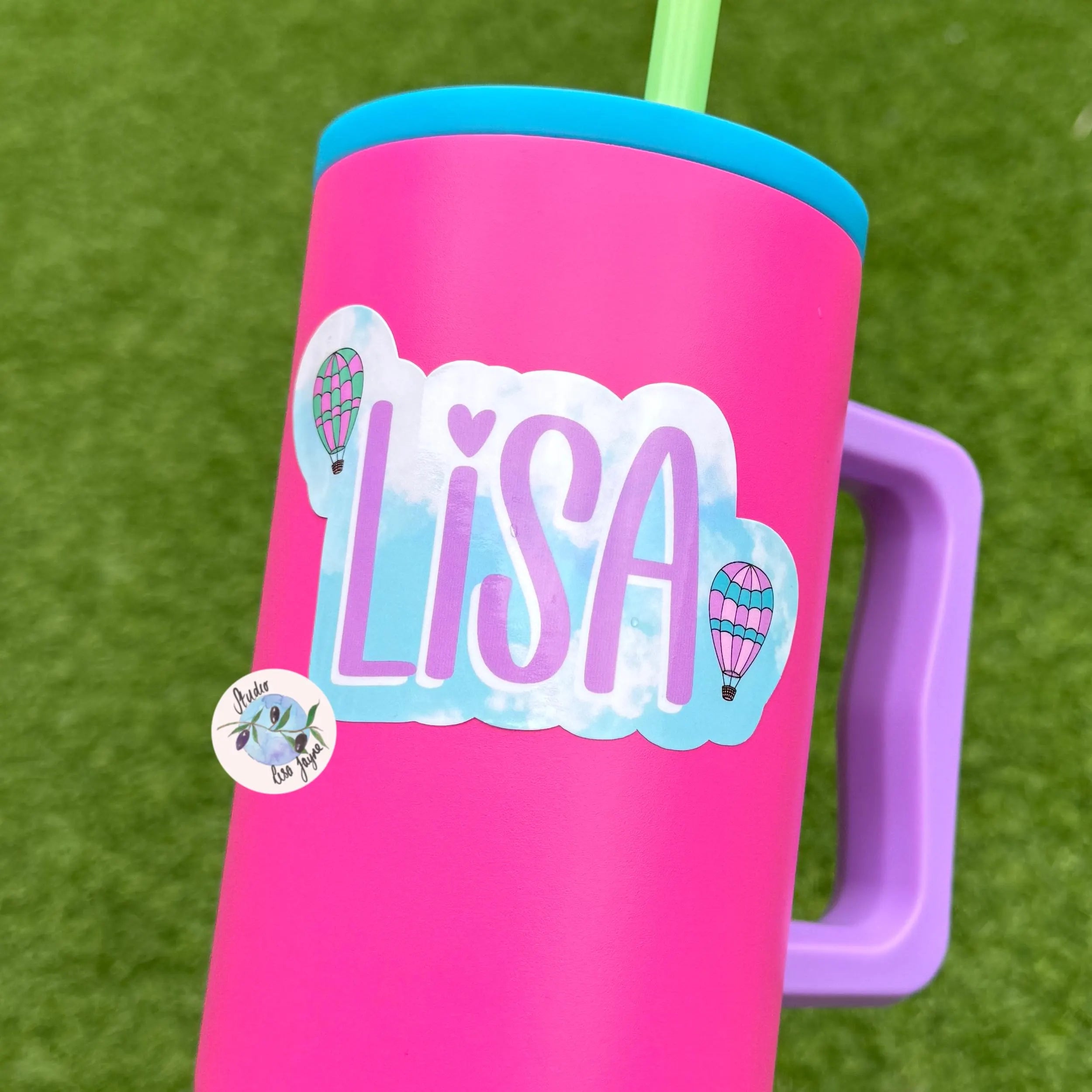 Pink tumbler with 'LISA' hot air baloon sticker against a grass background