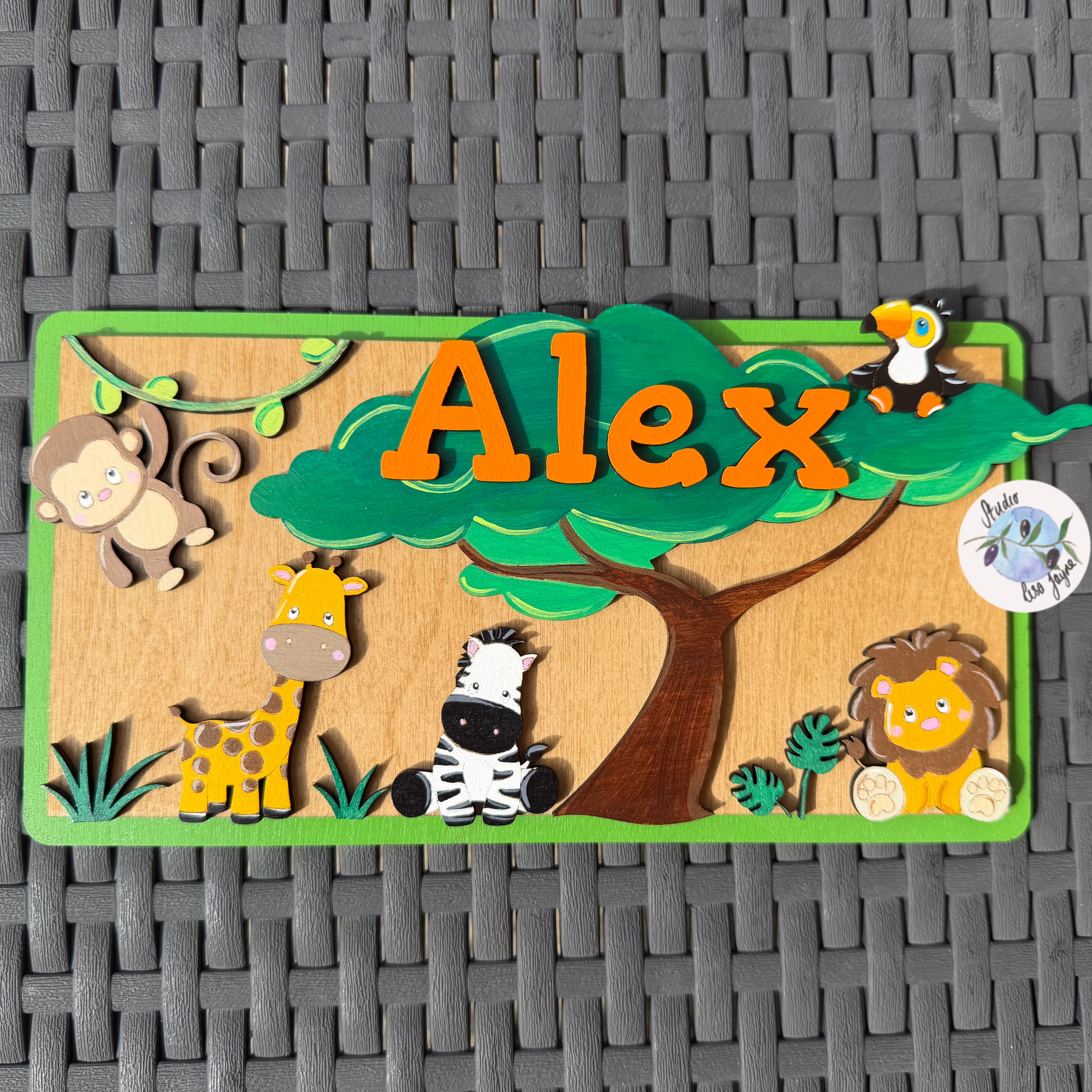 A personalized wooden name plaque featuring the name 'Alex' in orange letters under a green tree, surrounded by cartoon safari animals including a monkey, giraffe, zebra, lion, and penguin. The plaque is displayed on a gray woven mat background.
