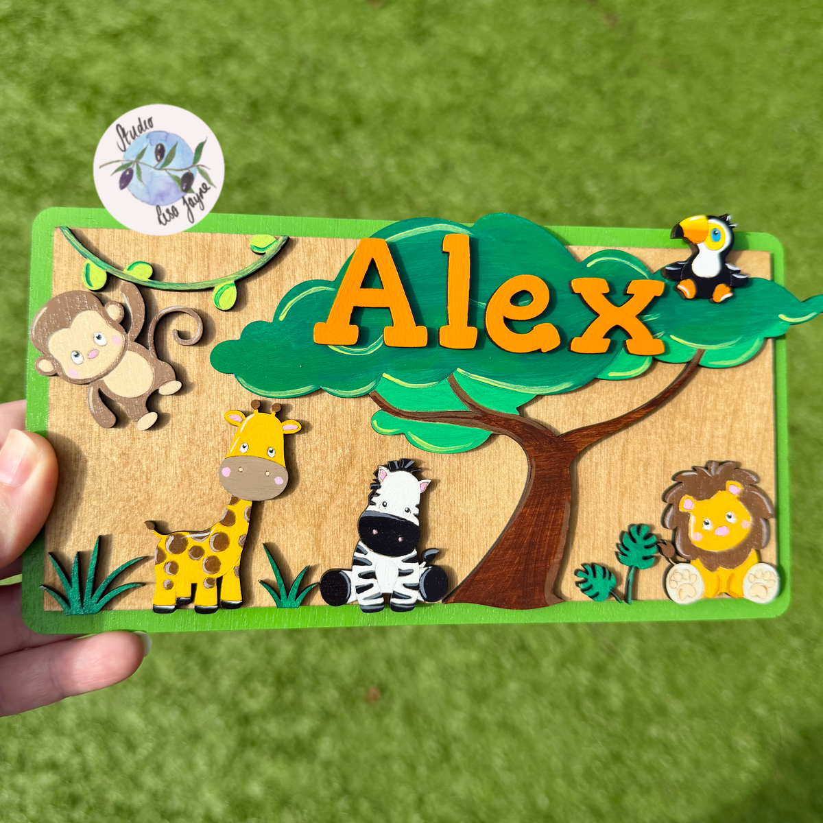 A personalized wooden name plaque featuring the name 'Alex' in orange letters under a green tree, surrounded by cartoon safari jungle animals including a monkey, giraffe, zebra, lion, and toucan. The plaque is held by a hand against a green grassy background.