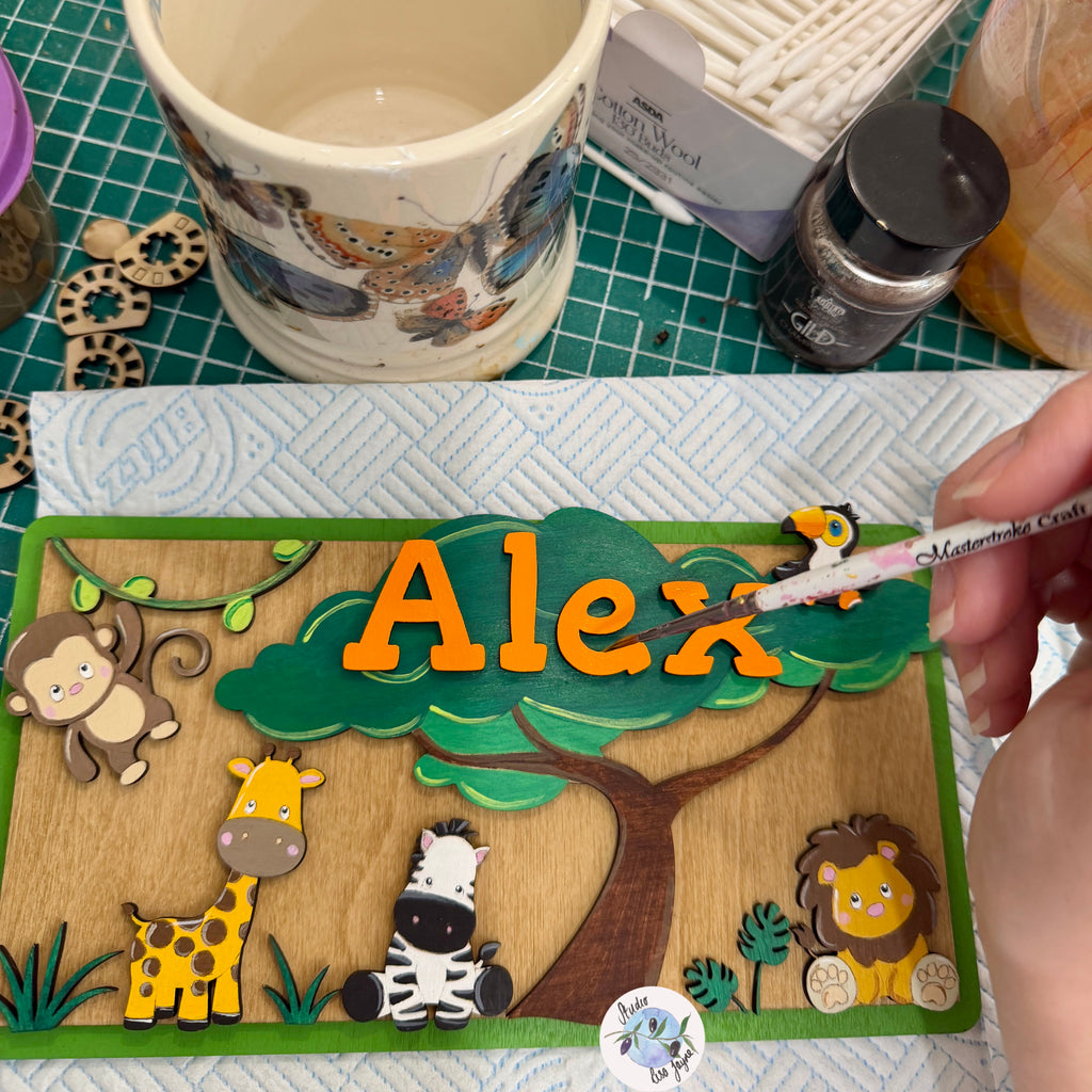 A close-up of a handmade wooden name plaque being painted. The plaque features a safari theme with a green tree, a brown trunk, and cartoon animals including a monkey, giraffe, zebra, and lion.