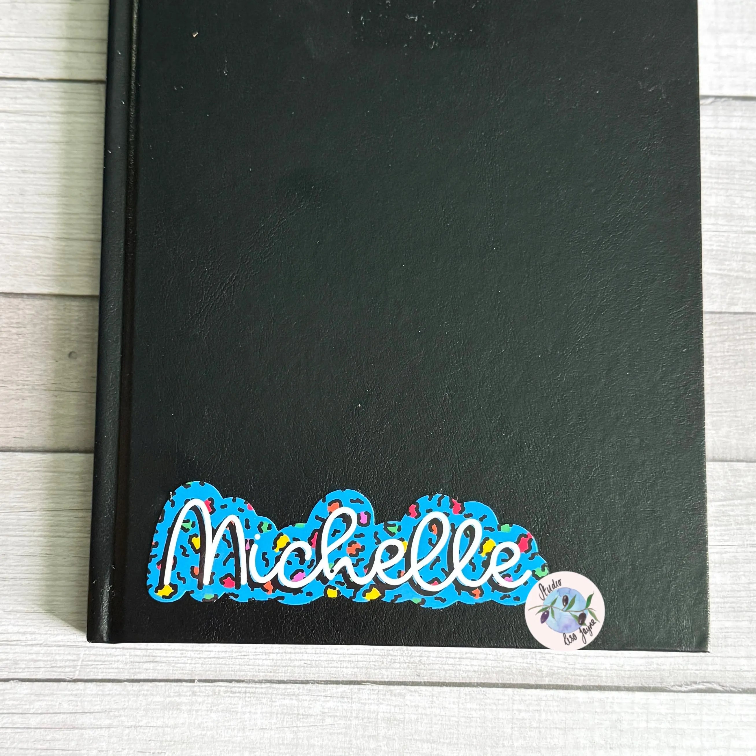 Black notebook with blue colorful leopard print name decal on a wooden surface
