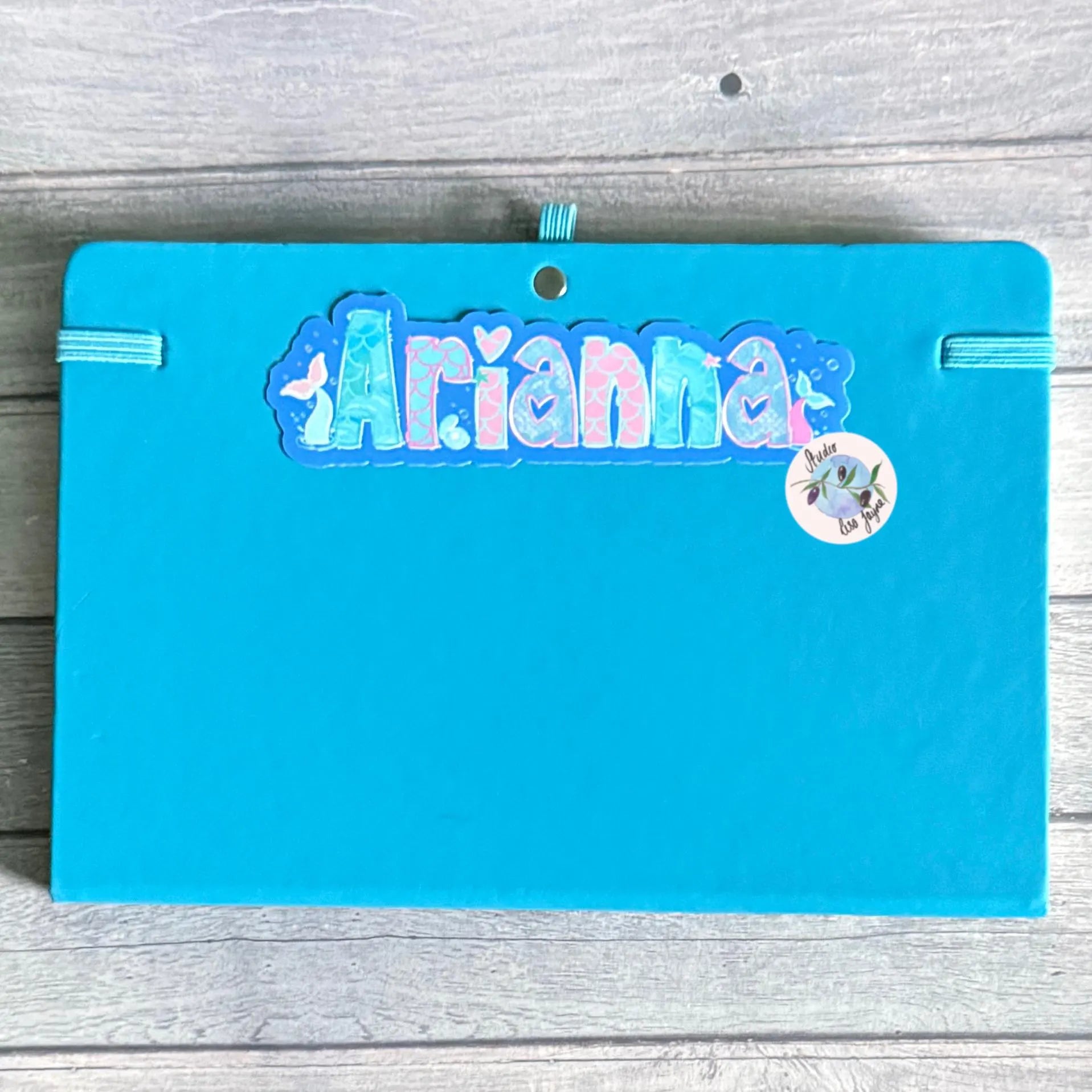 Blue notebook diary with 'Arianna' name sticker in a mermaid underwater theme on a wooden surface