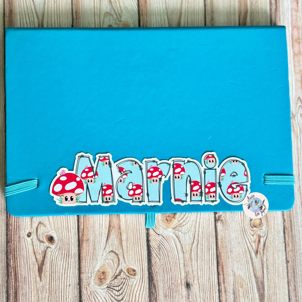 Blue notebook, diary with 'Marnie' and mushroom designs on a wooden surface