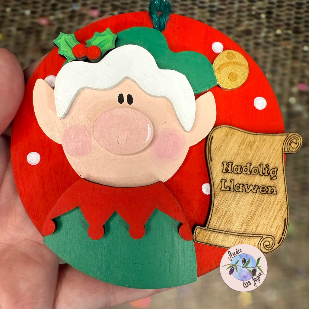 Nadolig Llawen Elf Christmas Tree Decoration Handmade Wooden Decoration Made in Wales - Green Elf on Red Backdrop in hand