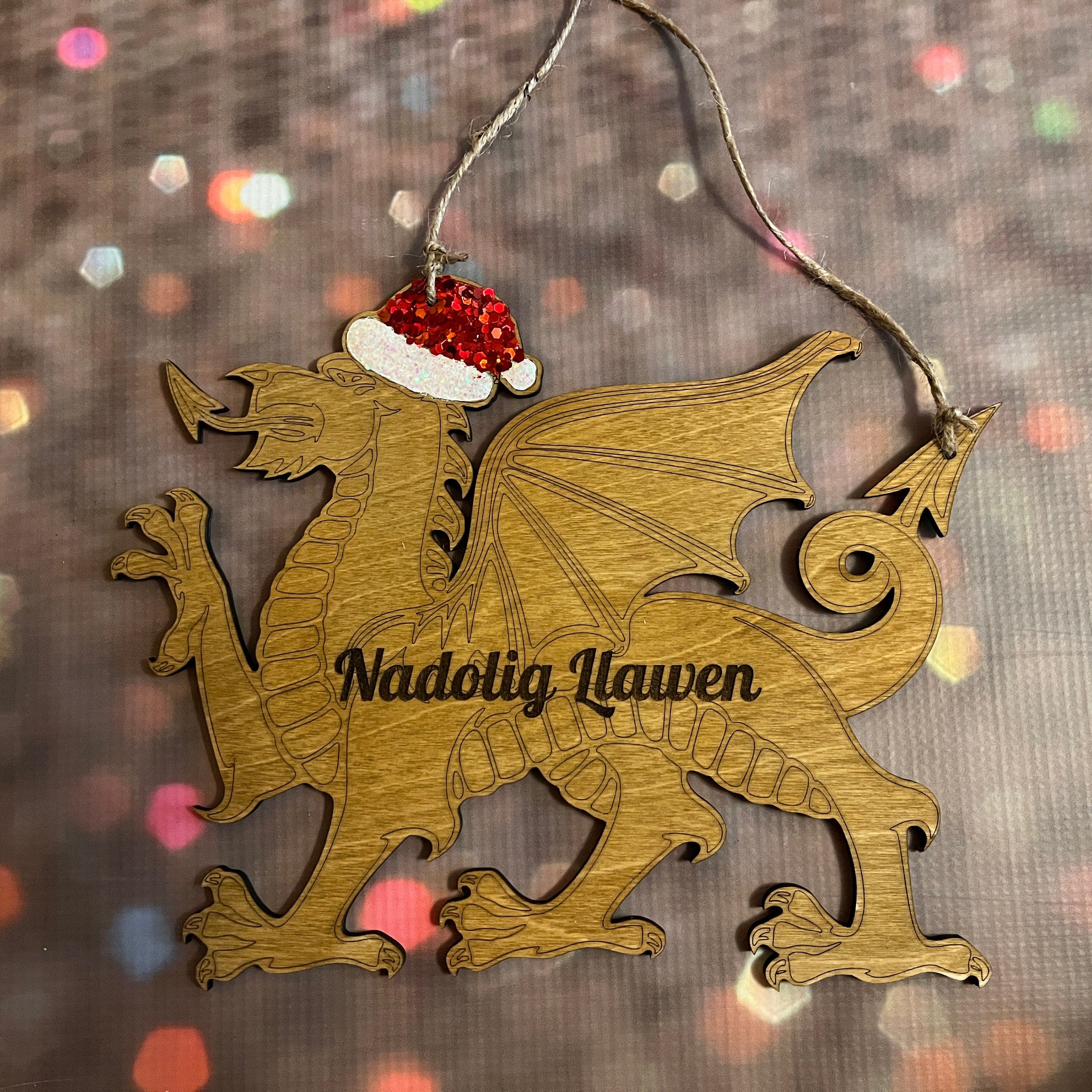 Wooden dragon ornament with a Santa hat and 'Nadolig Llawen' text on a blurred Christmas lights background.