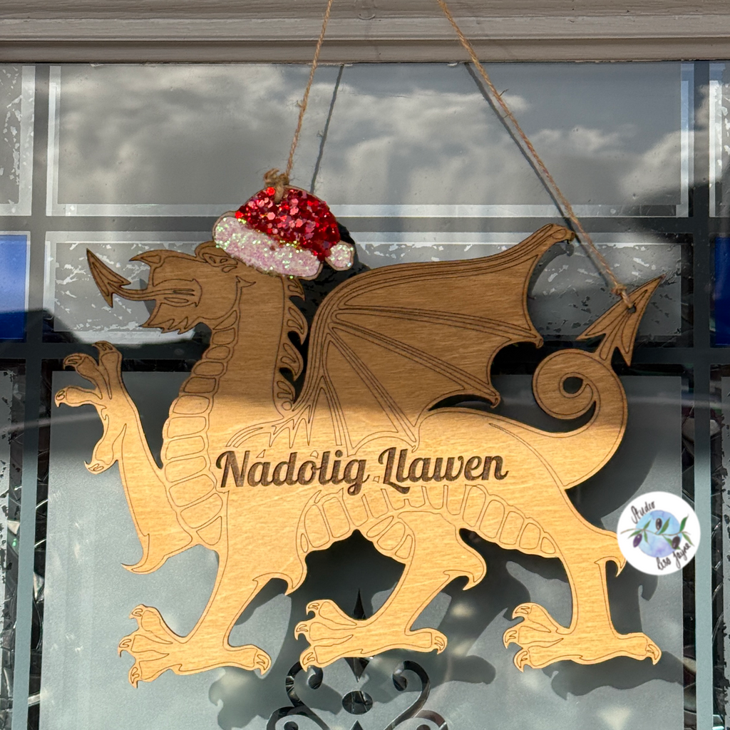 Wooden Welsh Christmas dragon ornament with a red and white hat, featuring the text 'Nadolig Llawen', against a glass window background.