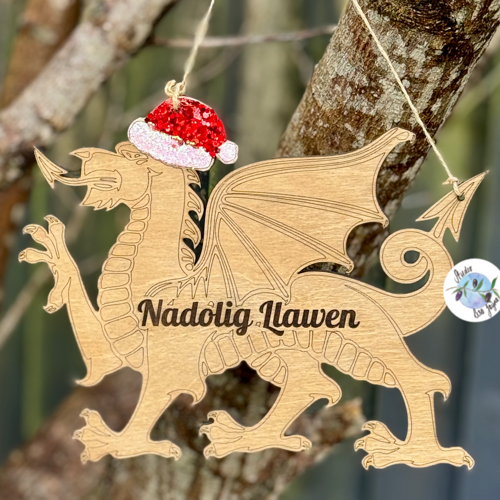 Wooden dragon hanging sign ornament with a glitter Santa hat hanging on a tree branch.  With the Welsh Christmas message Nadolg Llawen