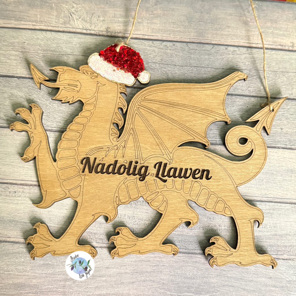 Wooden welsh dragon Christmas ornament with a Santa hat on a wooden surface.  Engraved Nadolig Llawen (Merry Christmas) message