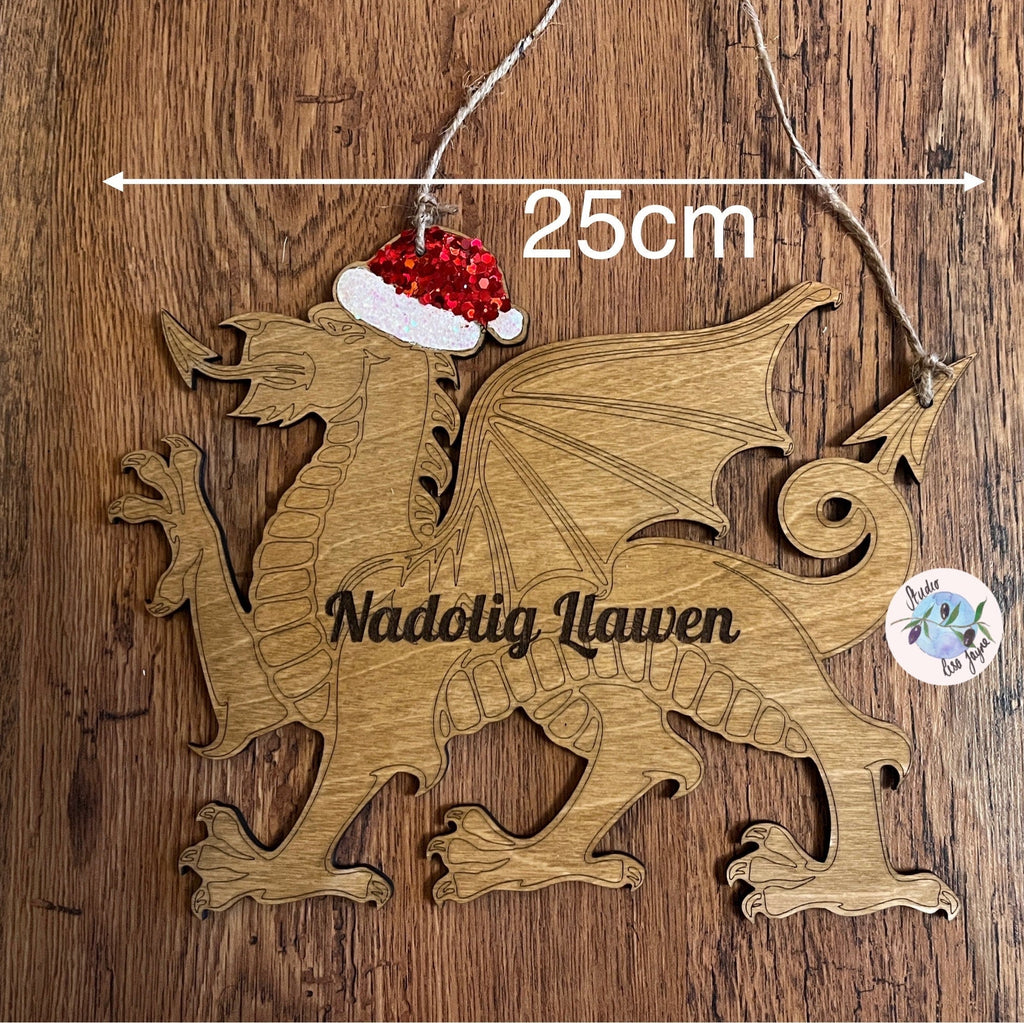 A wooden hanging sign featuring a red Welsh dragon with a Santa hat, the text 'Nadolig Llawen' carved in the center, and decorated with red and silver glitter. The sign is 25cm wide.